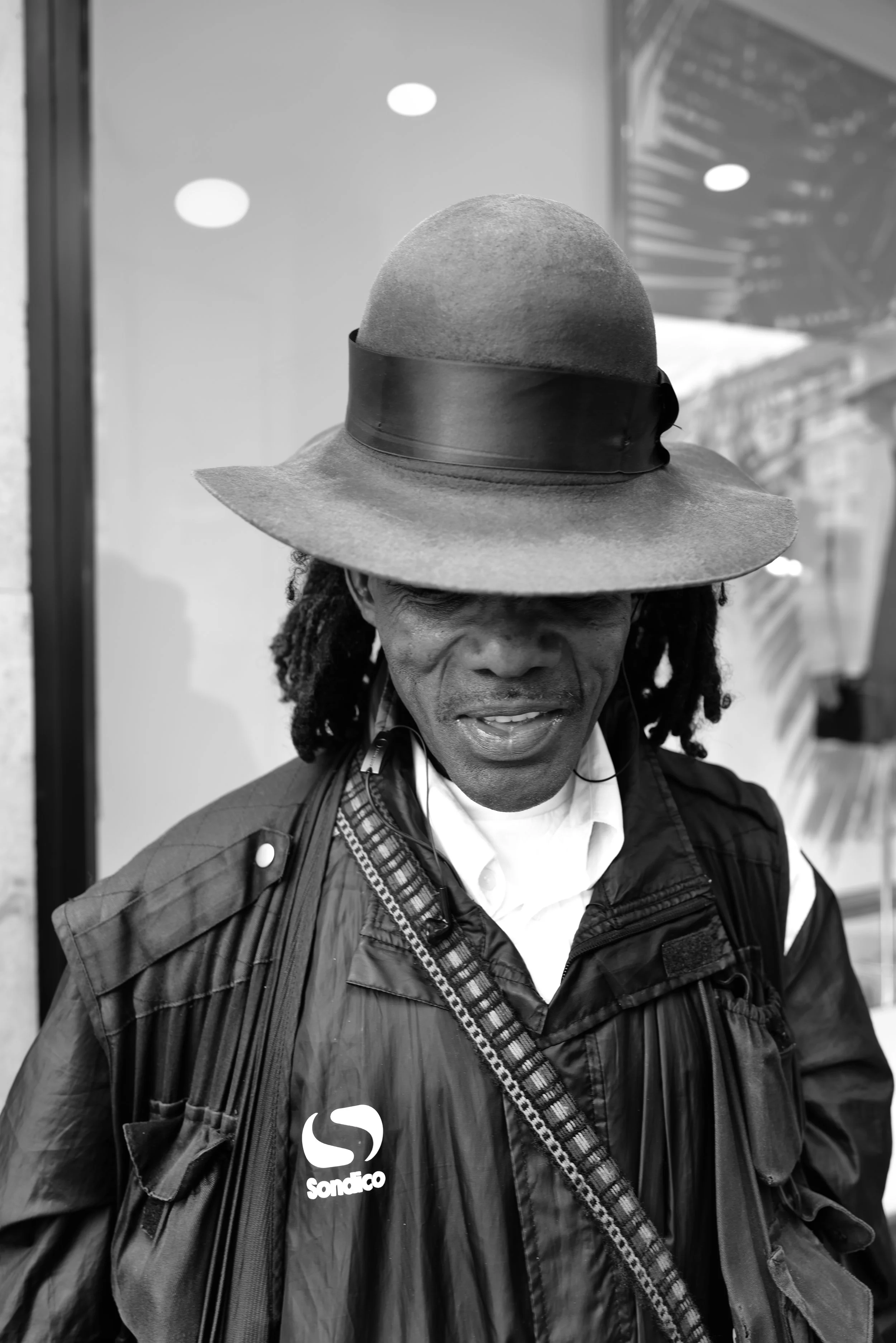 A man wearing a wide-brimmed hat, a jacket, and a shirt underneath, with a bag strap across his chest, standing indoors.