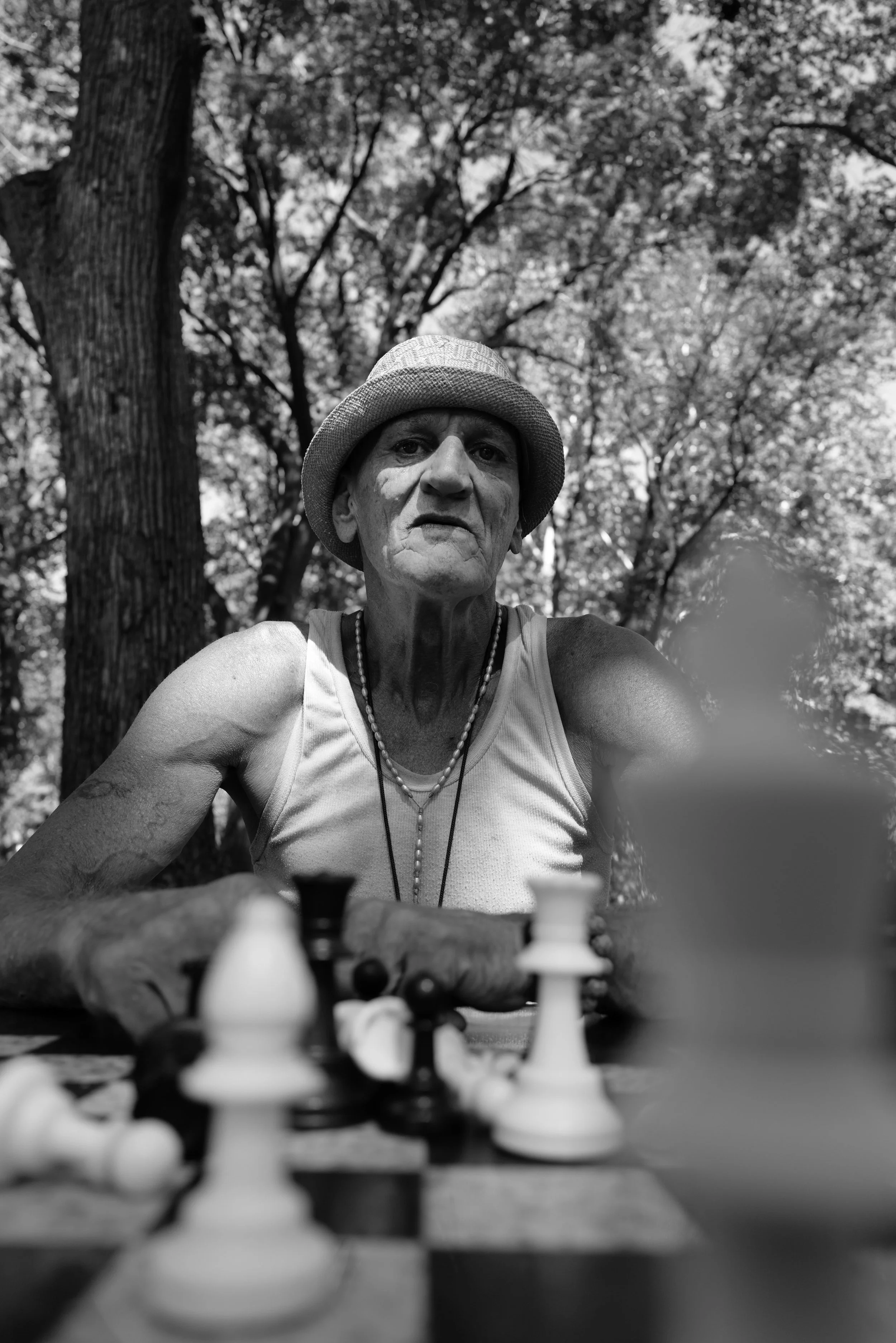An elderly man playing chess outdoors under a tree, wearing a hat and tank top, with a serious expression on his face.