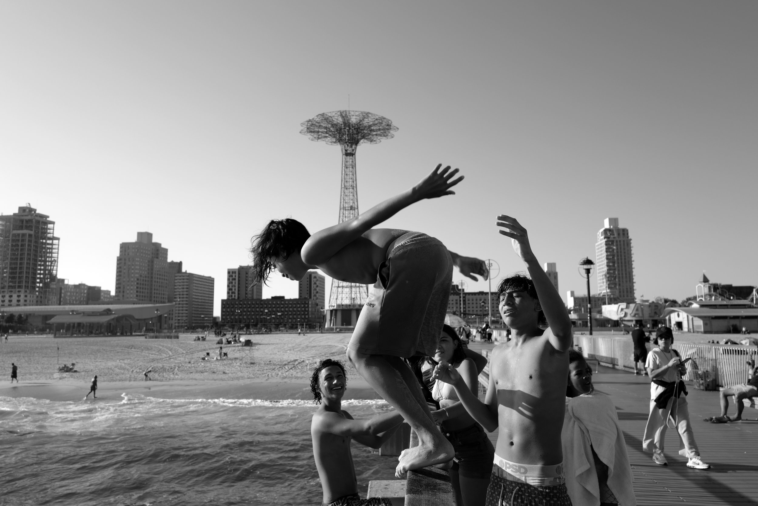 People jumping into the water at a beach, with a city skyline and a tall tower in the background.