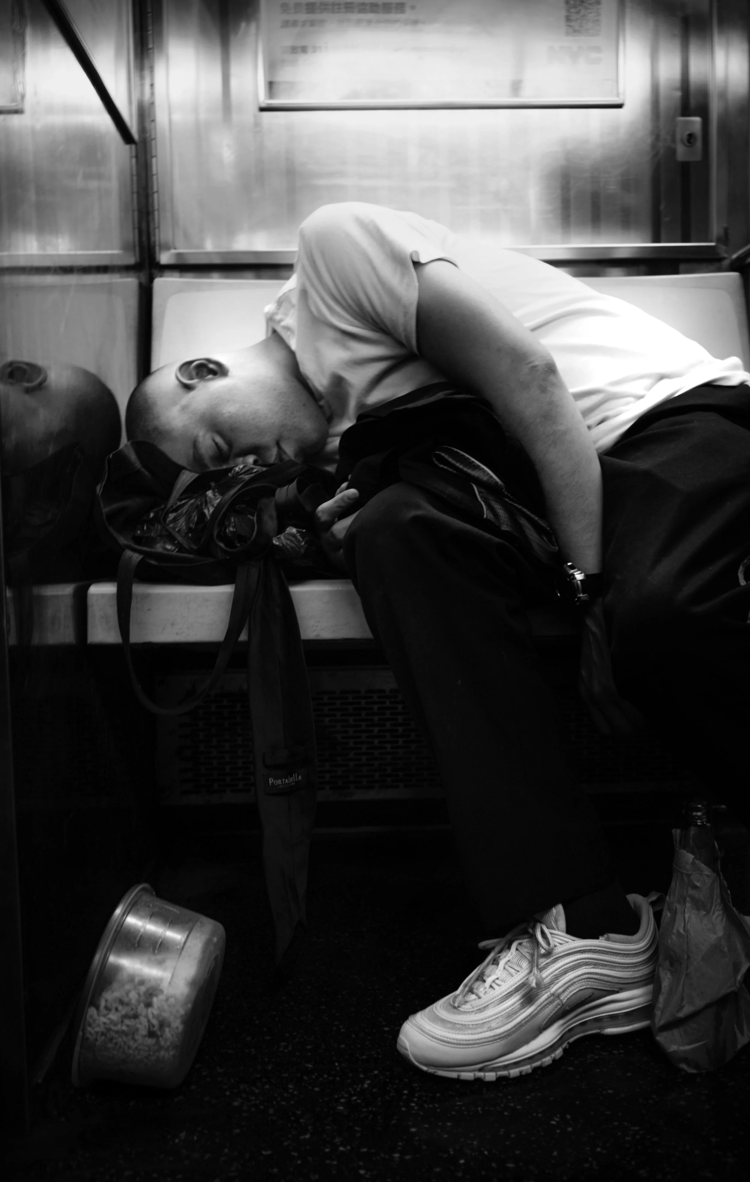 A man is sleeping on a train, lying on a bench with his head resting on a backpack, in a black and white photo.