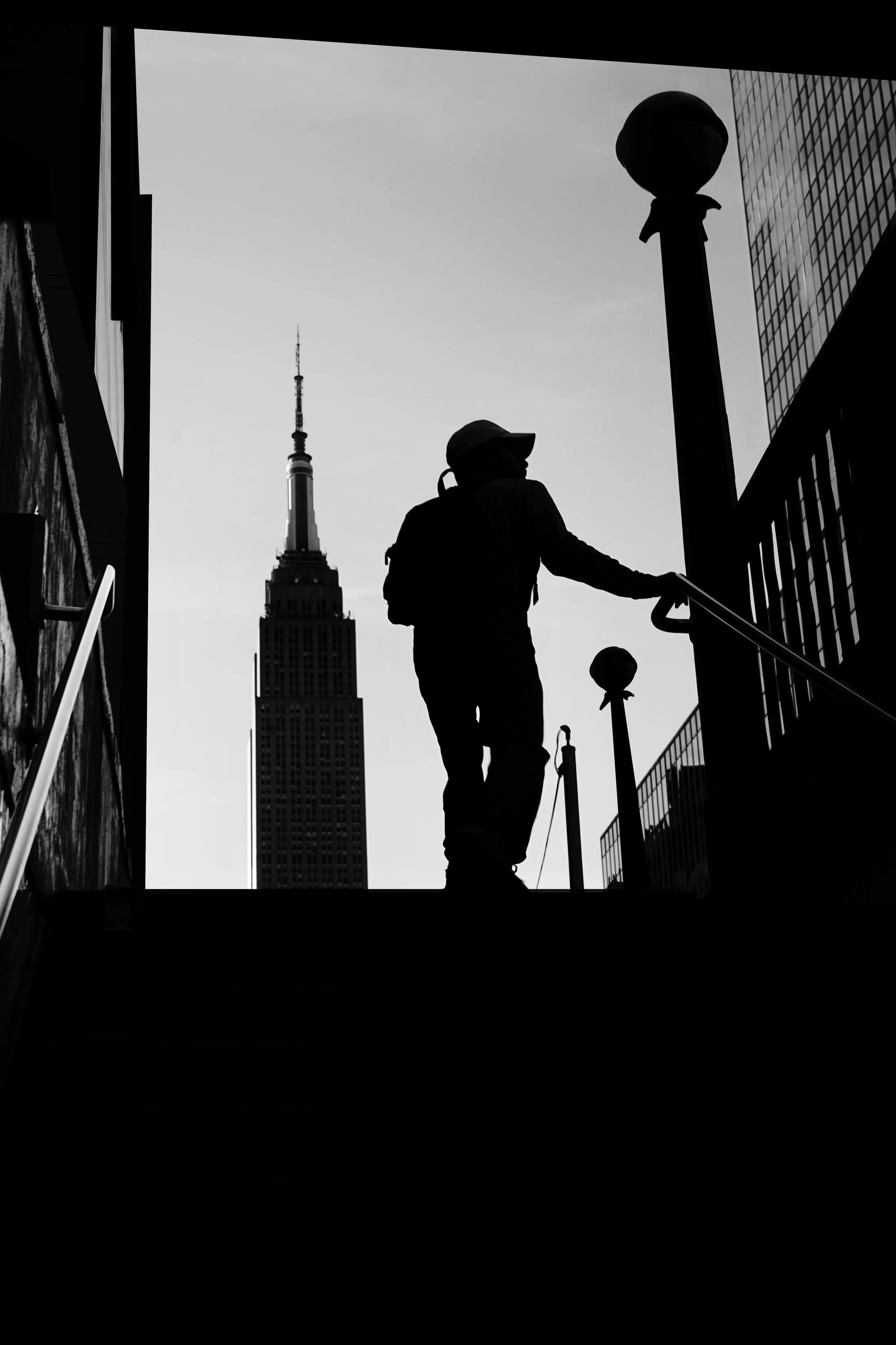 Silhouette of a person wearing a hat and headphones, walking on stairs near buildings in New York City, with the Empire State Building in the background.
