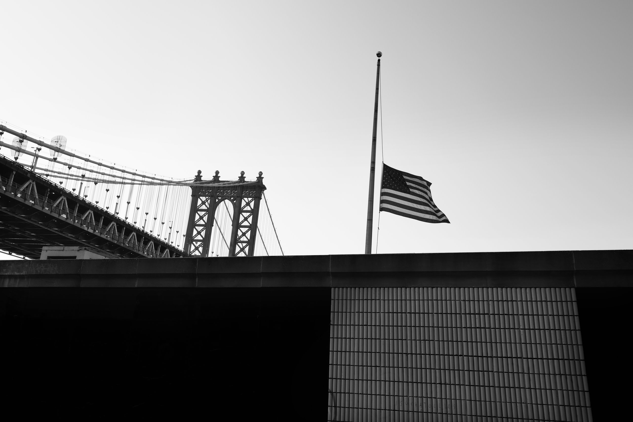 Black and white photo of an American flag on a pole in front of a modern building, with a bridge in the background.