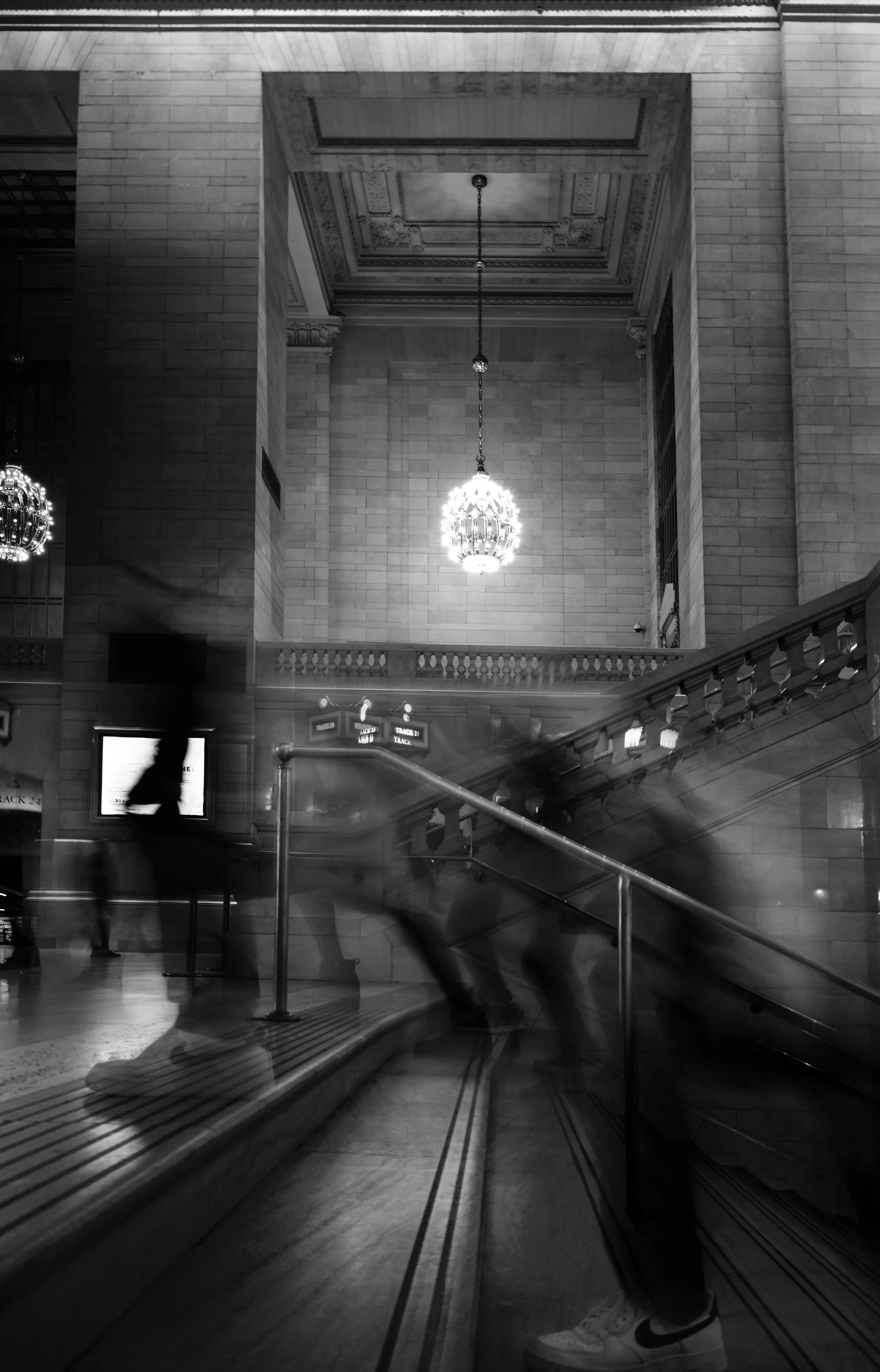 Interior of a grand building with high ceilings and ornate detailing, illuminated by hanging chandeliers, with blurred motion of people moving on a staircase.