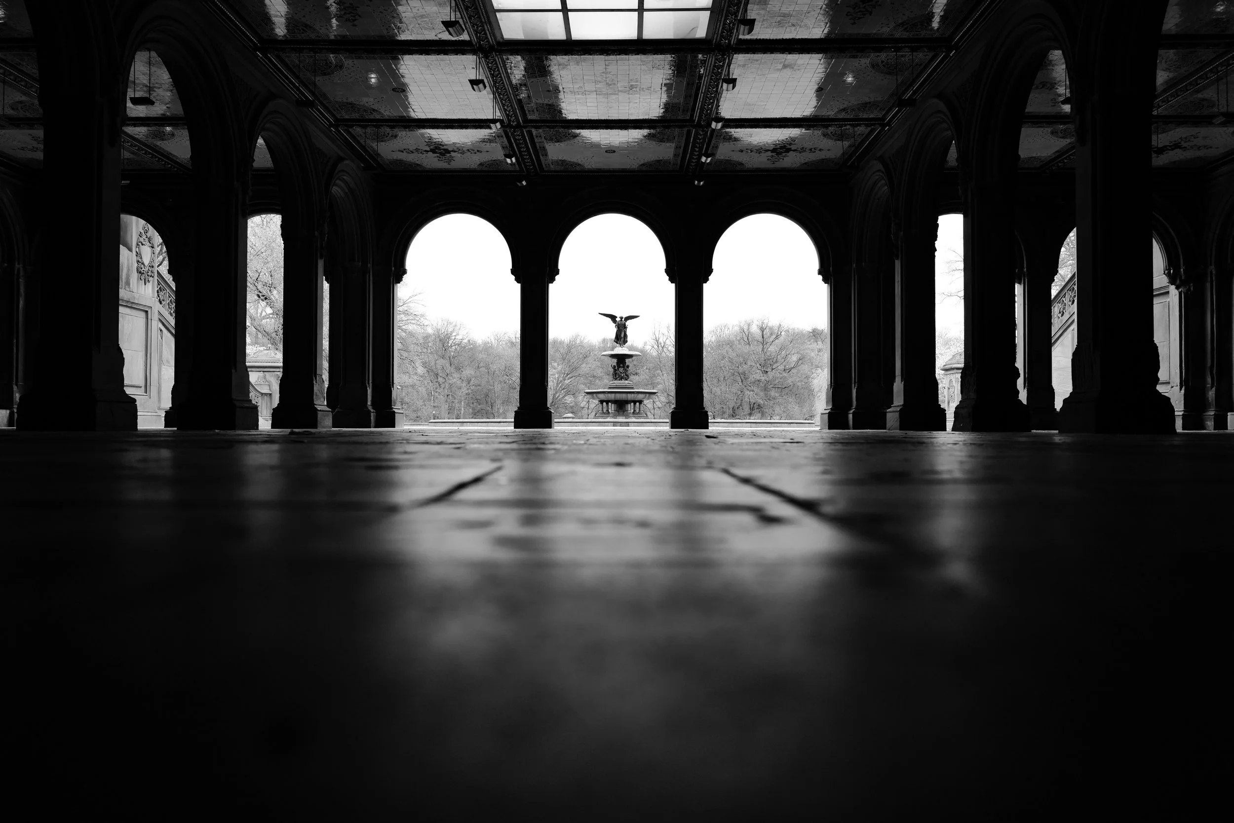 View of a decorative fountain through arched openings in a structure, with trees visible in the background, black and white photography.