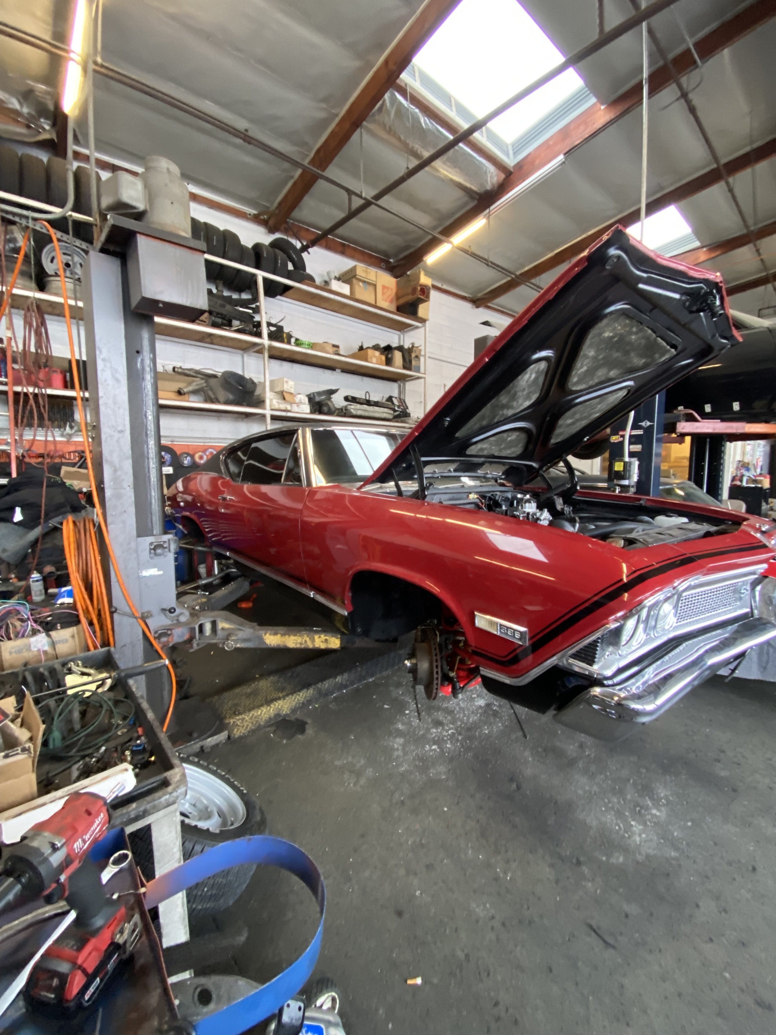 A red classic car with its hood open and front wheel removed in a workshop or garage.
