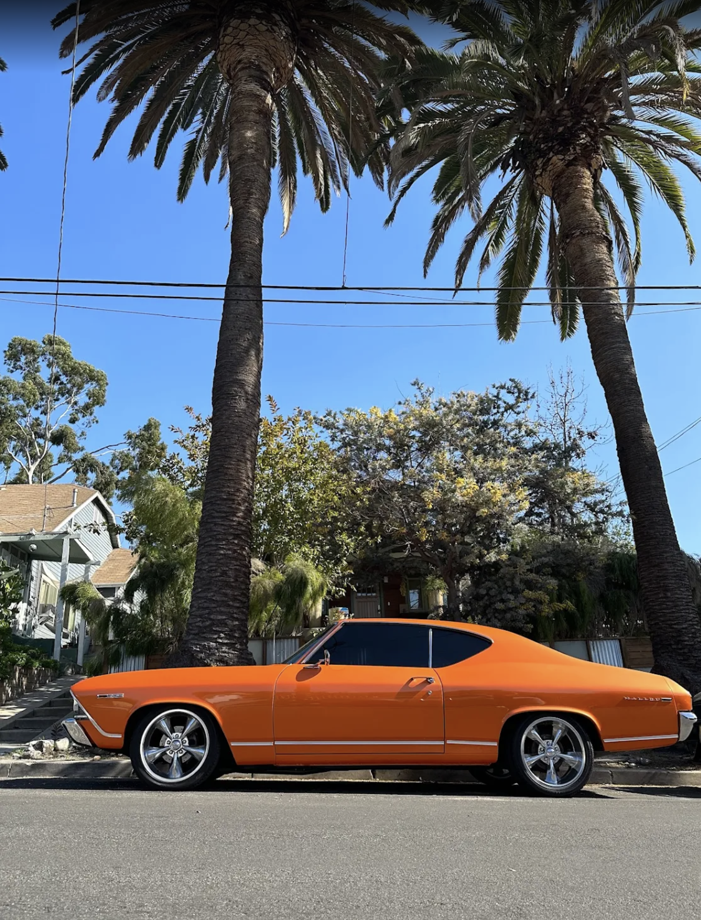 A vintage orange car parked on the street with two tall palm trees and a neighborhood with houses and trees in the background.