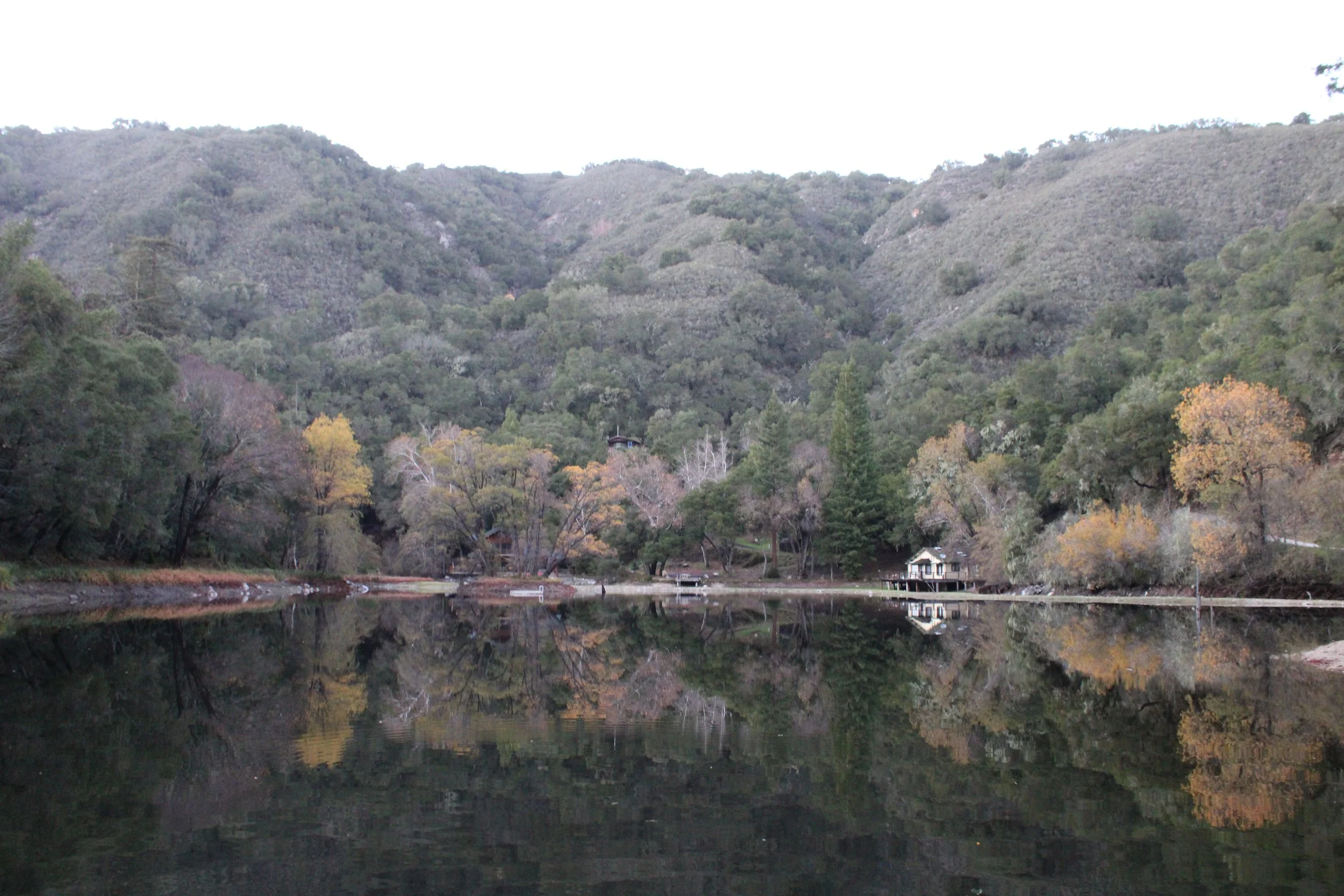 A calm lake reflecting trees and mountains in the background, with some trees displaying autumn colors and a small white house on the right side of the lake.