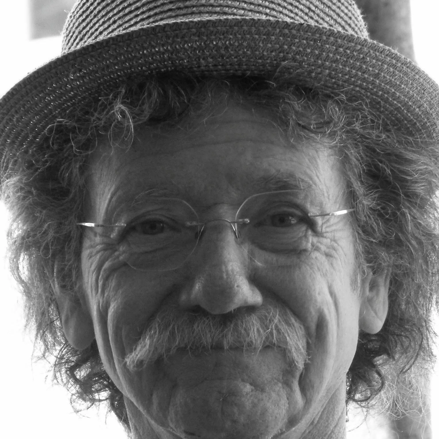 Close-up black and white photo of an older man with curly hair, glasses, a mustache and beard, wearing a straw hat.