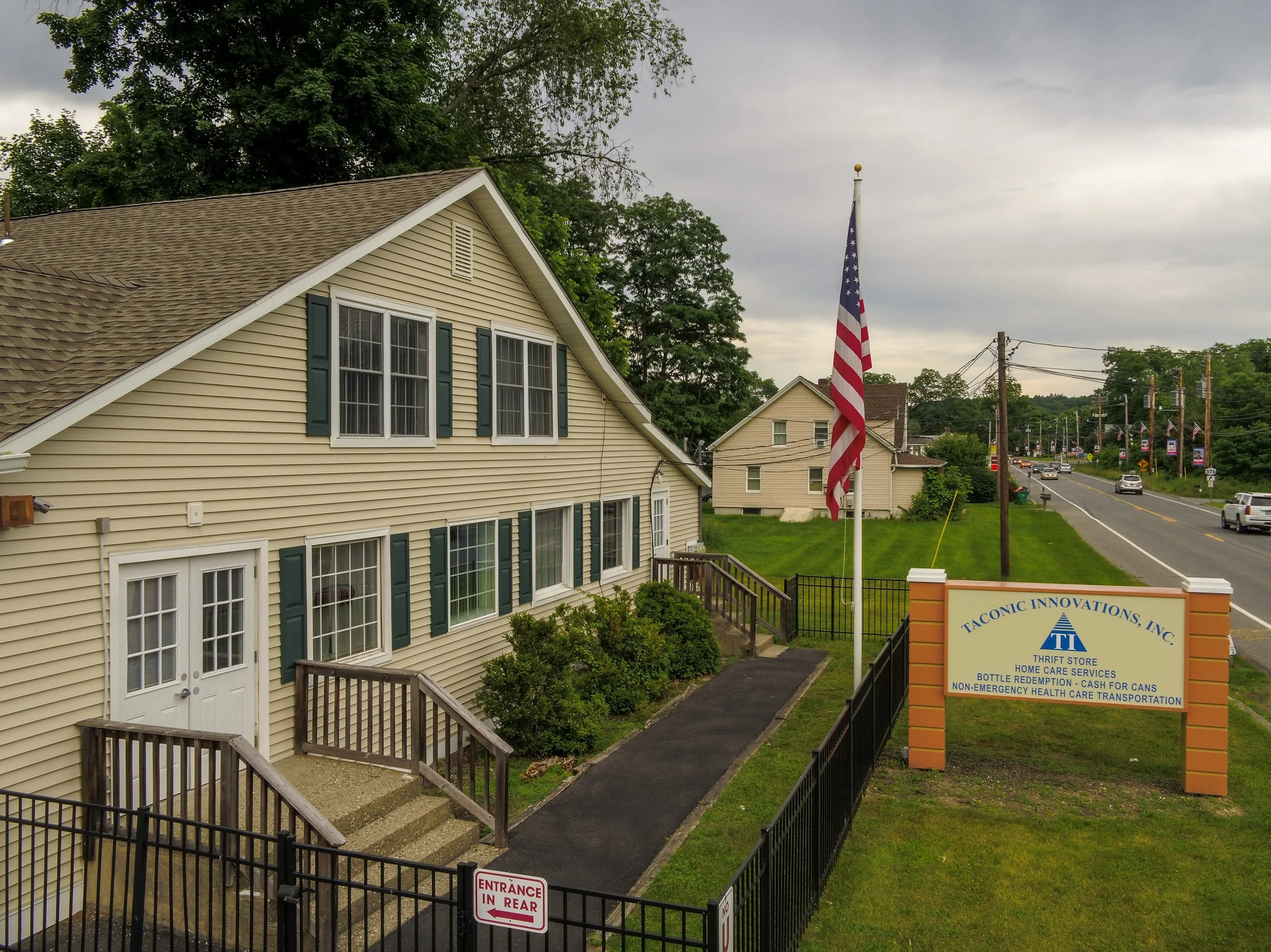 A yellow two-story building with green shutters and a white door, a sidewalk leading to the door, and a flagpole with an American flag outside. There is a sign for Taconic Innovations, Inc., and a road with cars in the background. The sky is overcast.
