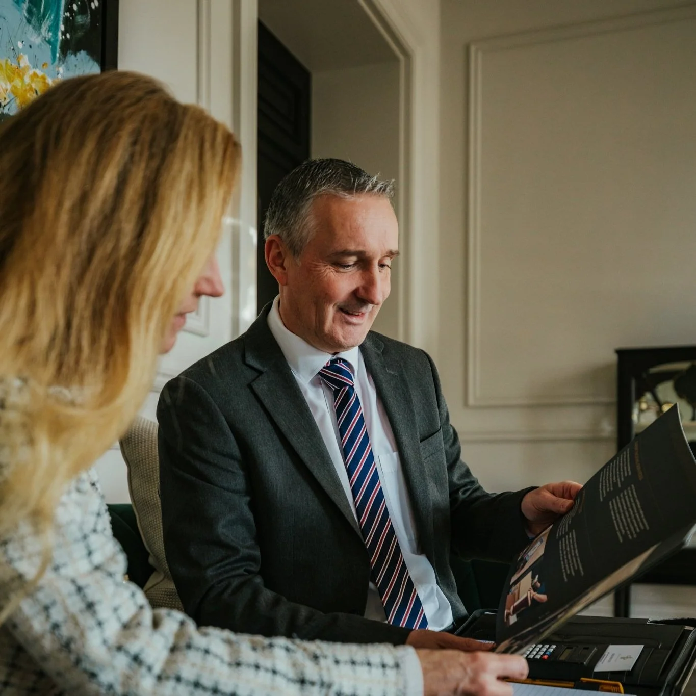 A man and a woman seated at a table, looking at a booklet or magazine together, in a well-lit room with beige walls and framed artwork.