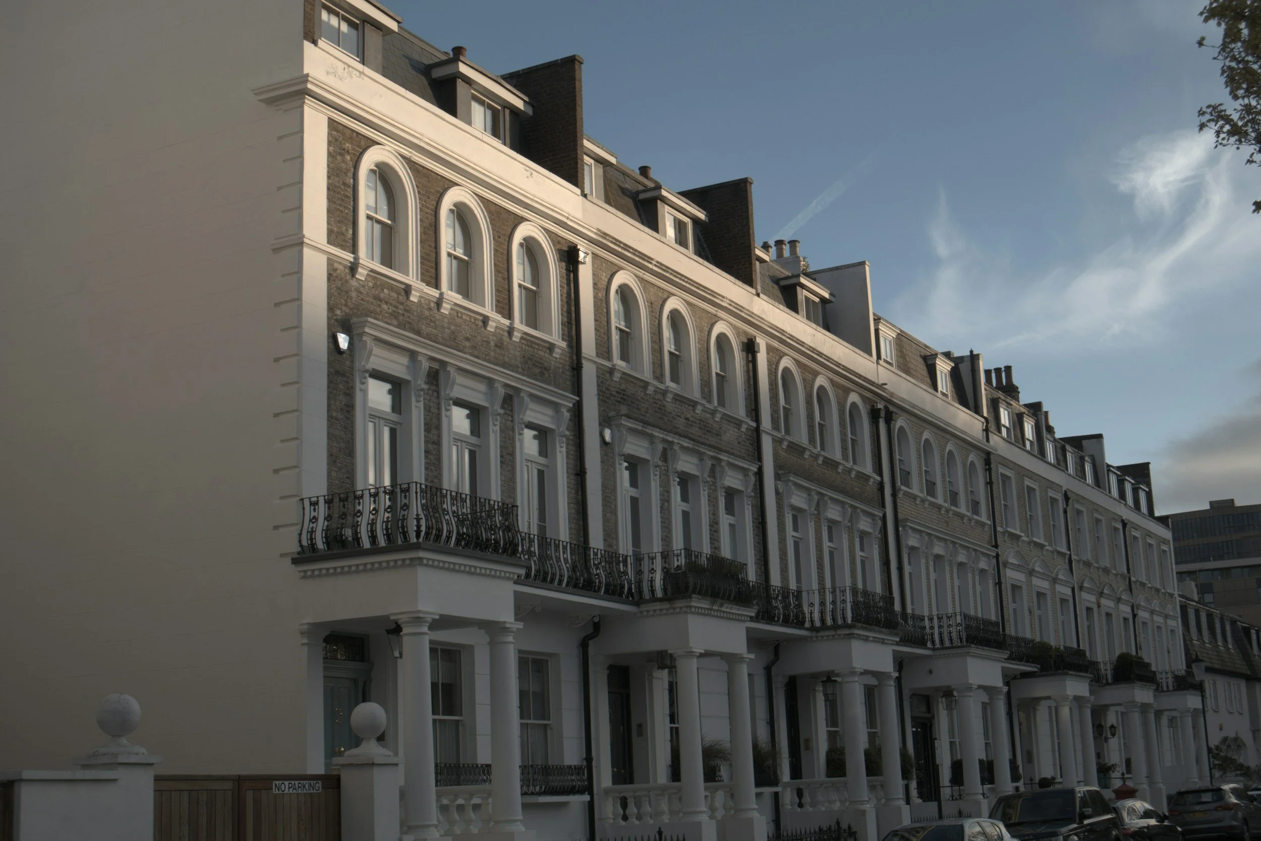A row of multi-story residential buildings with white and brick exteriors, arched windows, and balconies, under a partly cloudy sky.