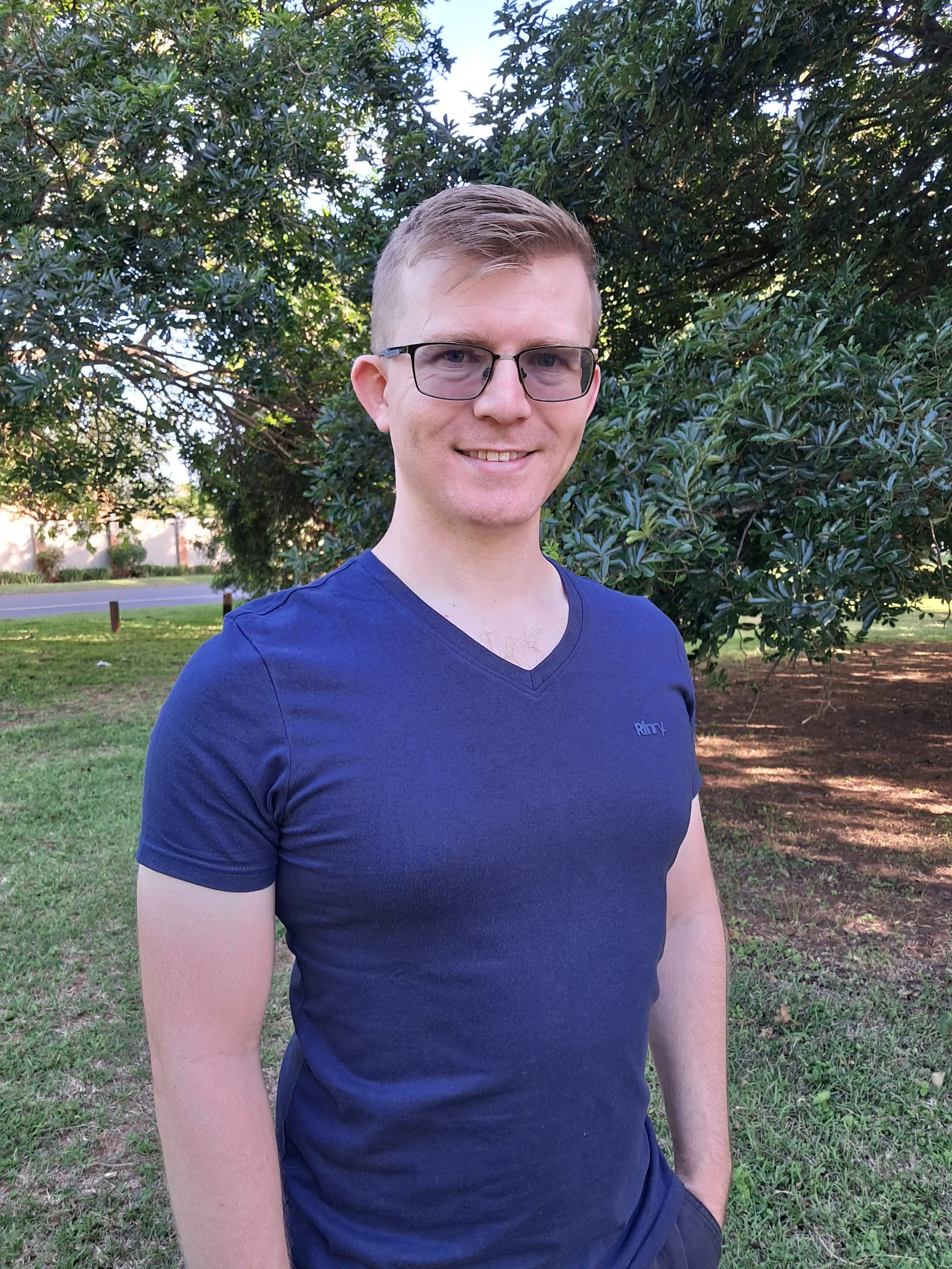 A young man with short blond hair, glasses, wearing a blue V-neck T-shirt, standing outdoors in a park with green grass and trees in the background, smiling at the camera.