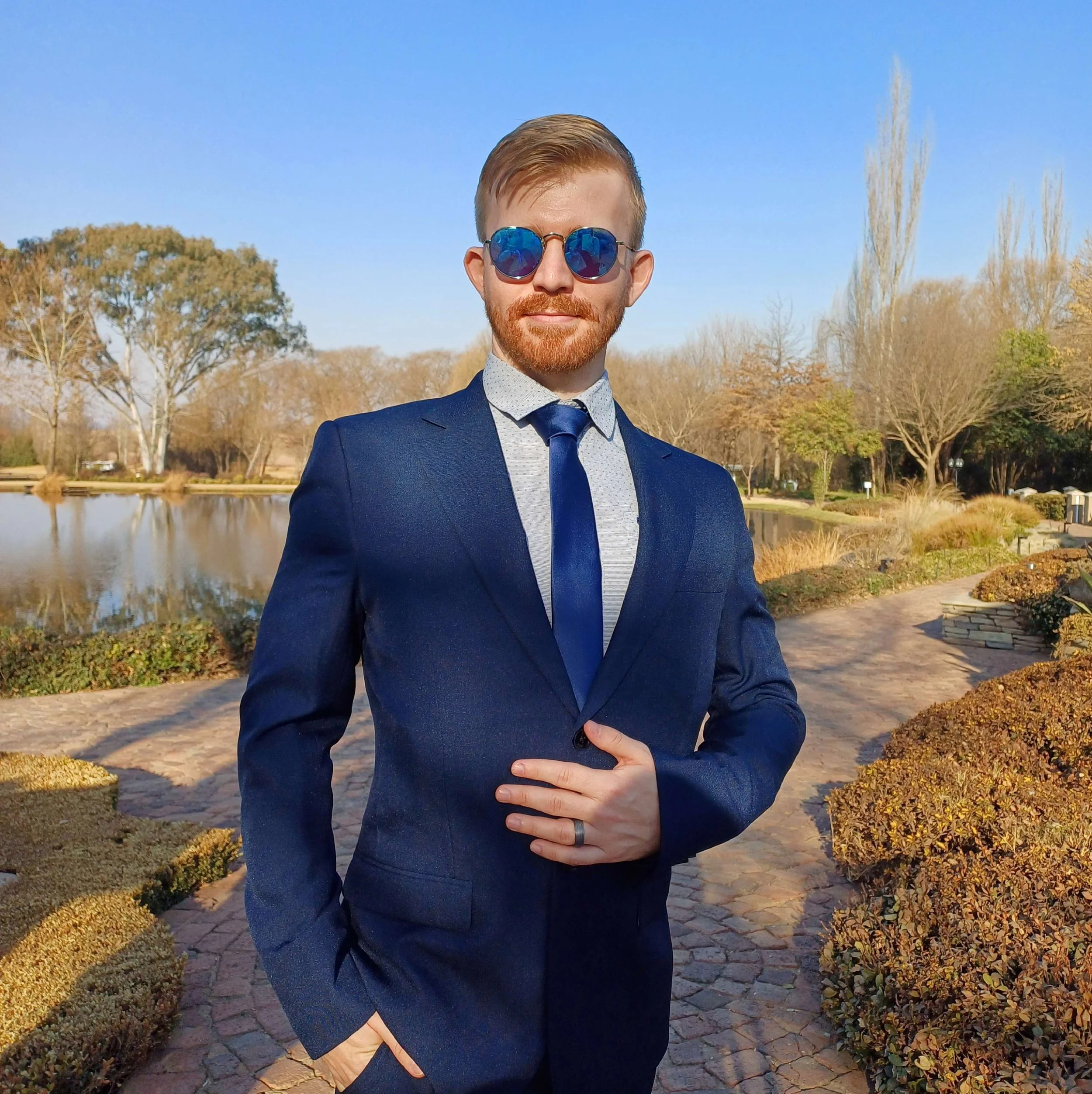 Man in a navy blue suit with a tie, wearing mirrored sunglasses, standing outdoors in front of a pond and trees on a clear sunny day.