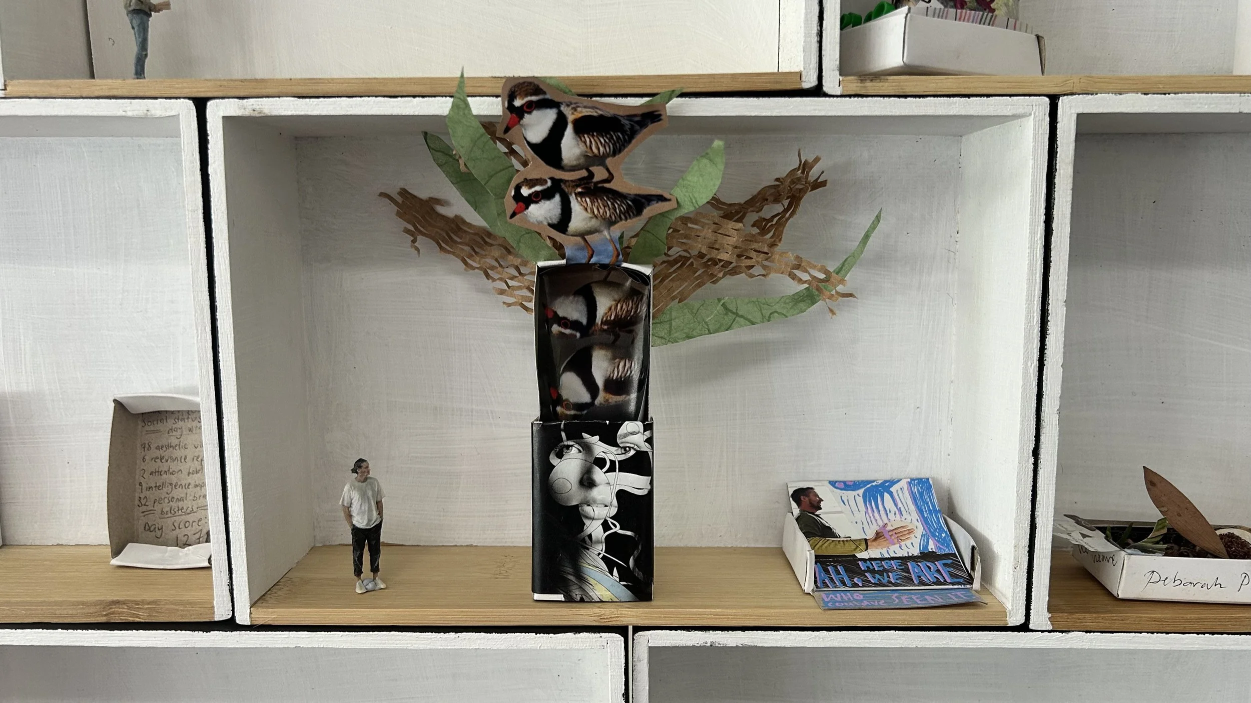 Decorative display with paper birds perched on a feed tube, surrounded by paper leaves and branches, set against a white box shelf with small figurines and items.