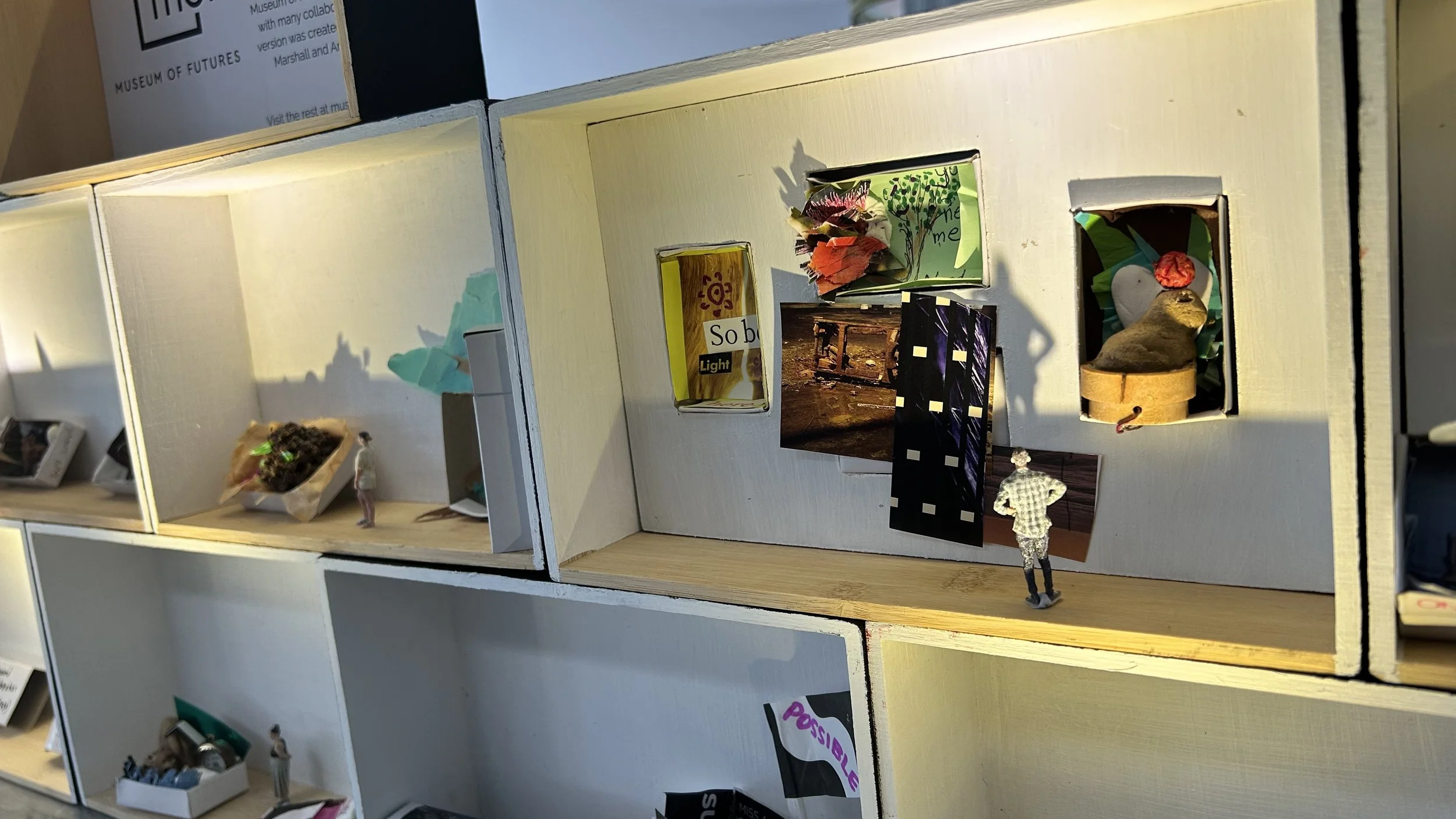 Miniature display cases with small art pieces and figurines, some with paper or fabric decorations.