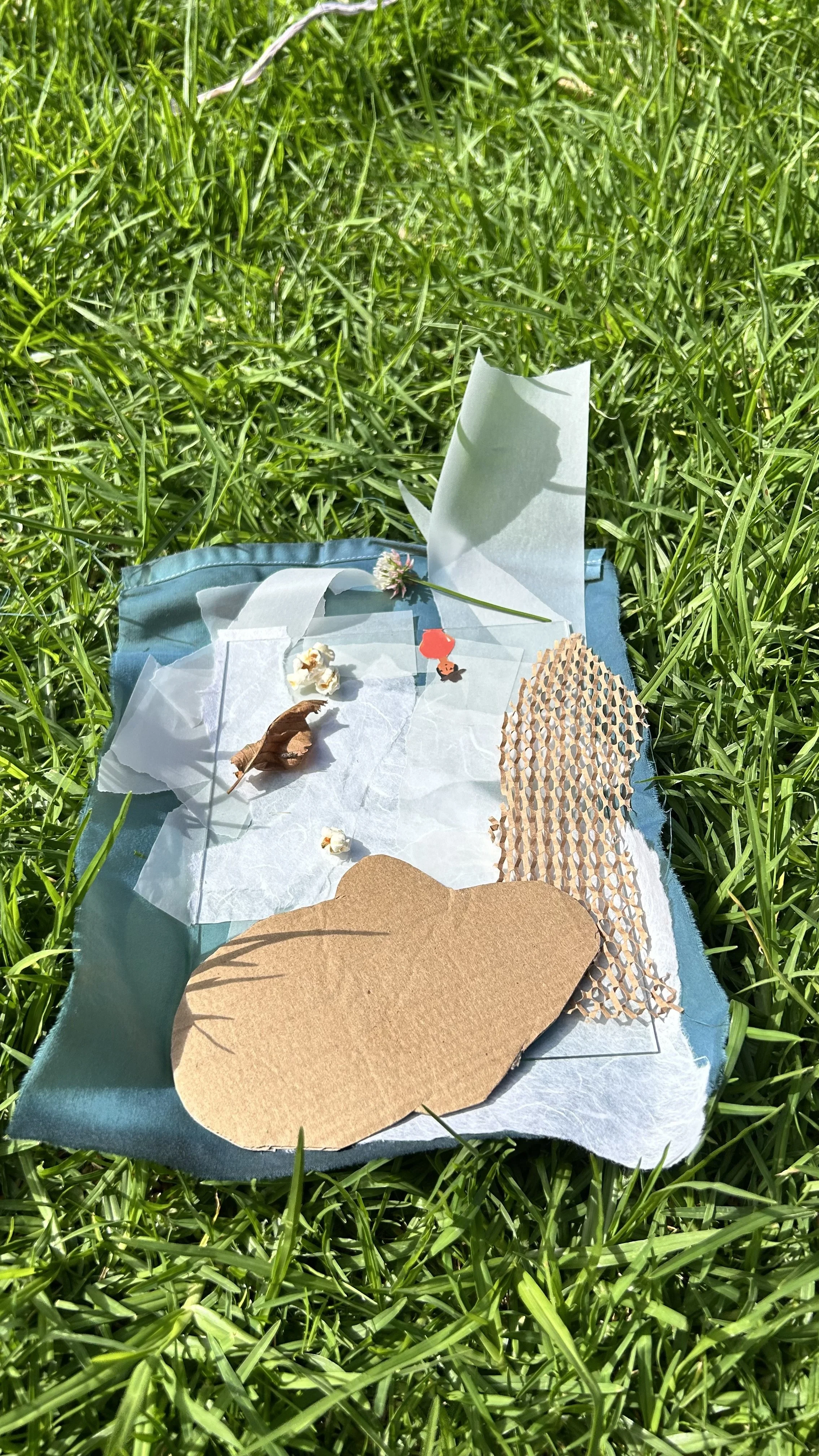 A small art installation on a piece of light blue fabric on green grass, featuring paper cutouts, a small flower, a popcorn. and a tiny figurine with a red umbrella.
