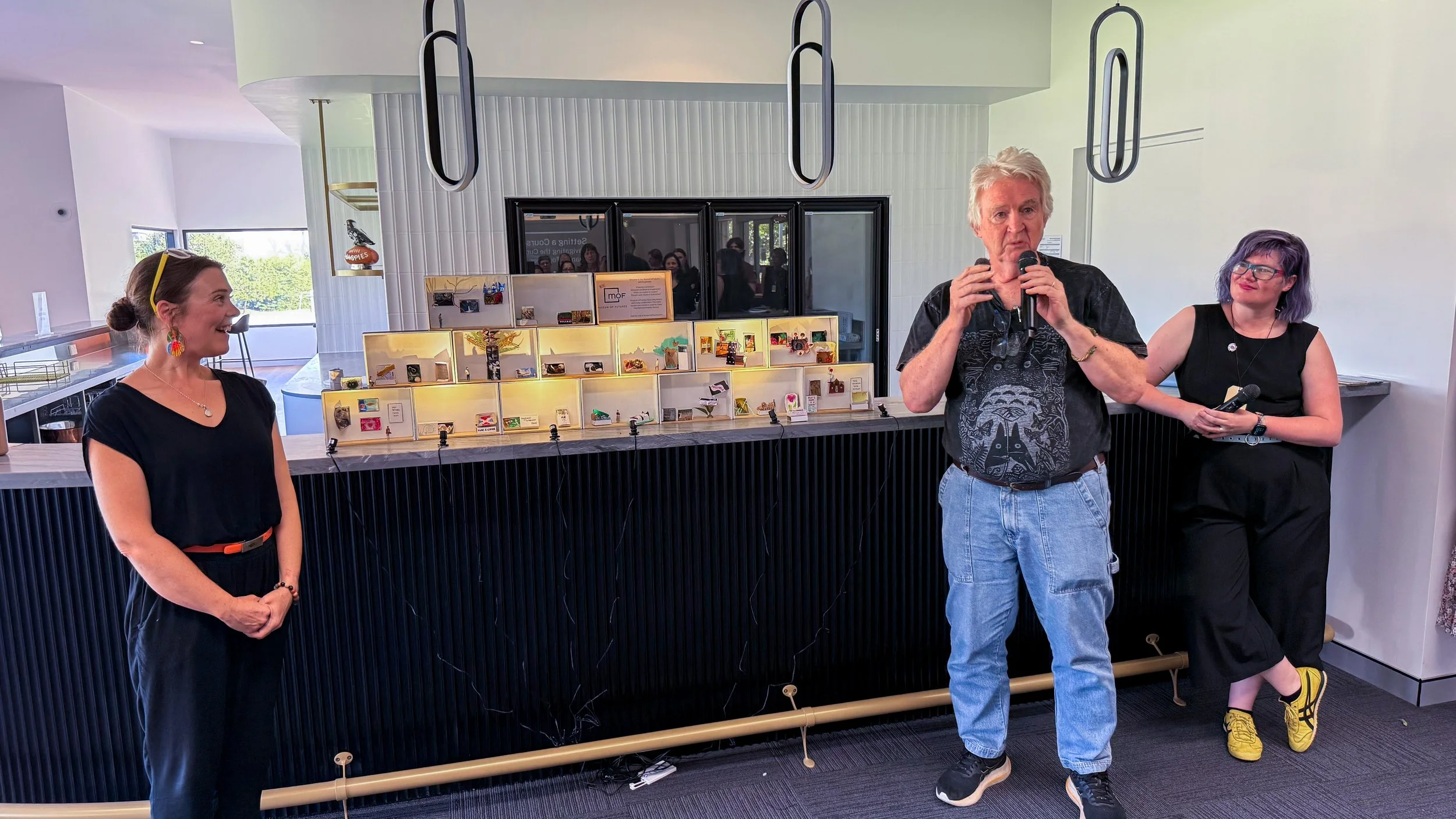 Three people standing in front of a display case filled with small items at an indoor event. The woman on the left is smiling and wearing a black outfit with colorful earrings. The man in the middle has white hair, is holding a microphone, and appears to be speaking. The woman on the right has short purple hair, glasses, and is holding a microphone, leaning against the wall.