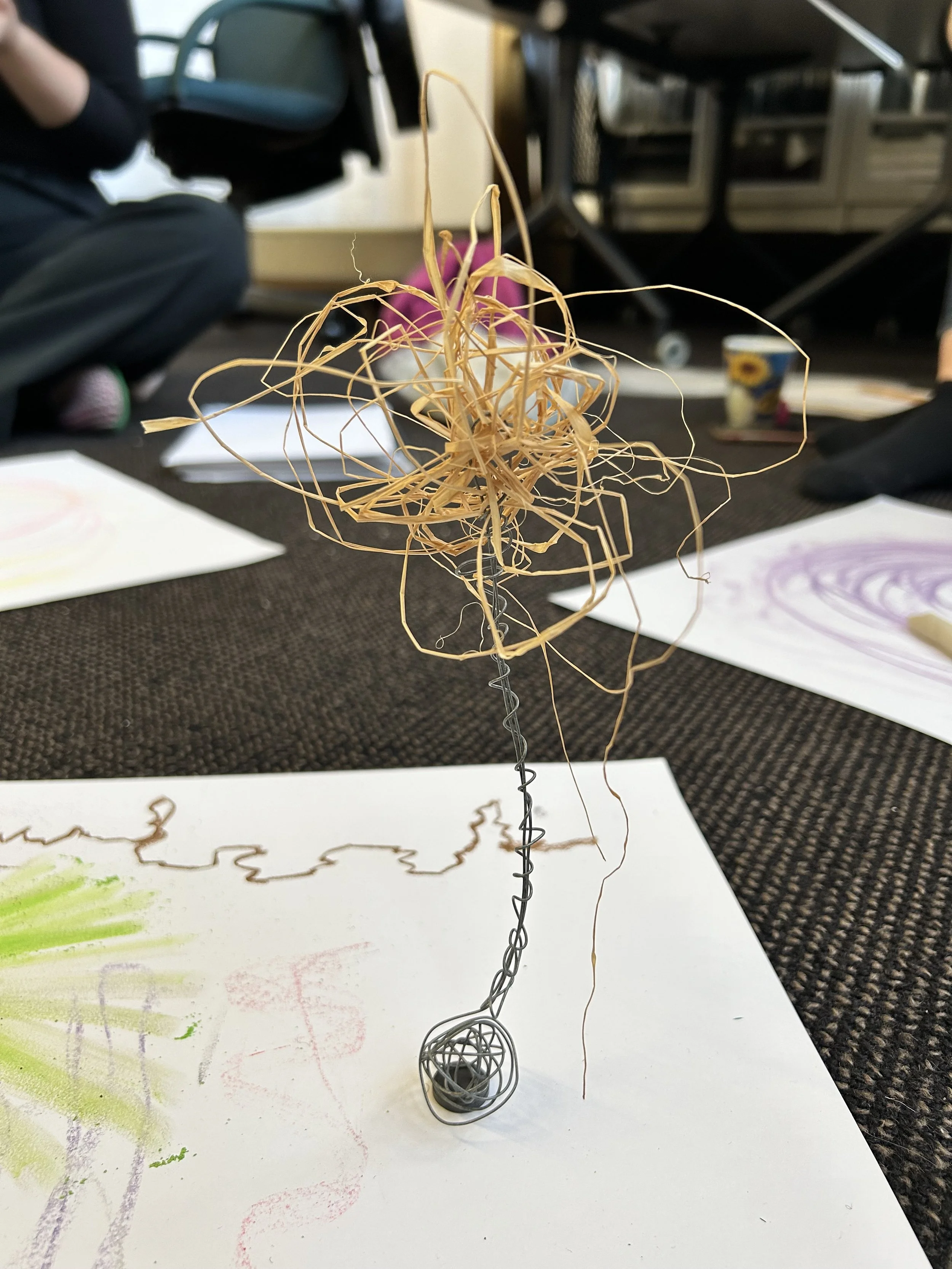 A wire sculpture with a tangled, abstract, and intricate design, standing on a black surface, with drawing papers and colorful drawings nearby.