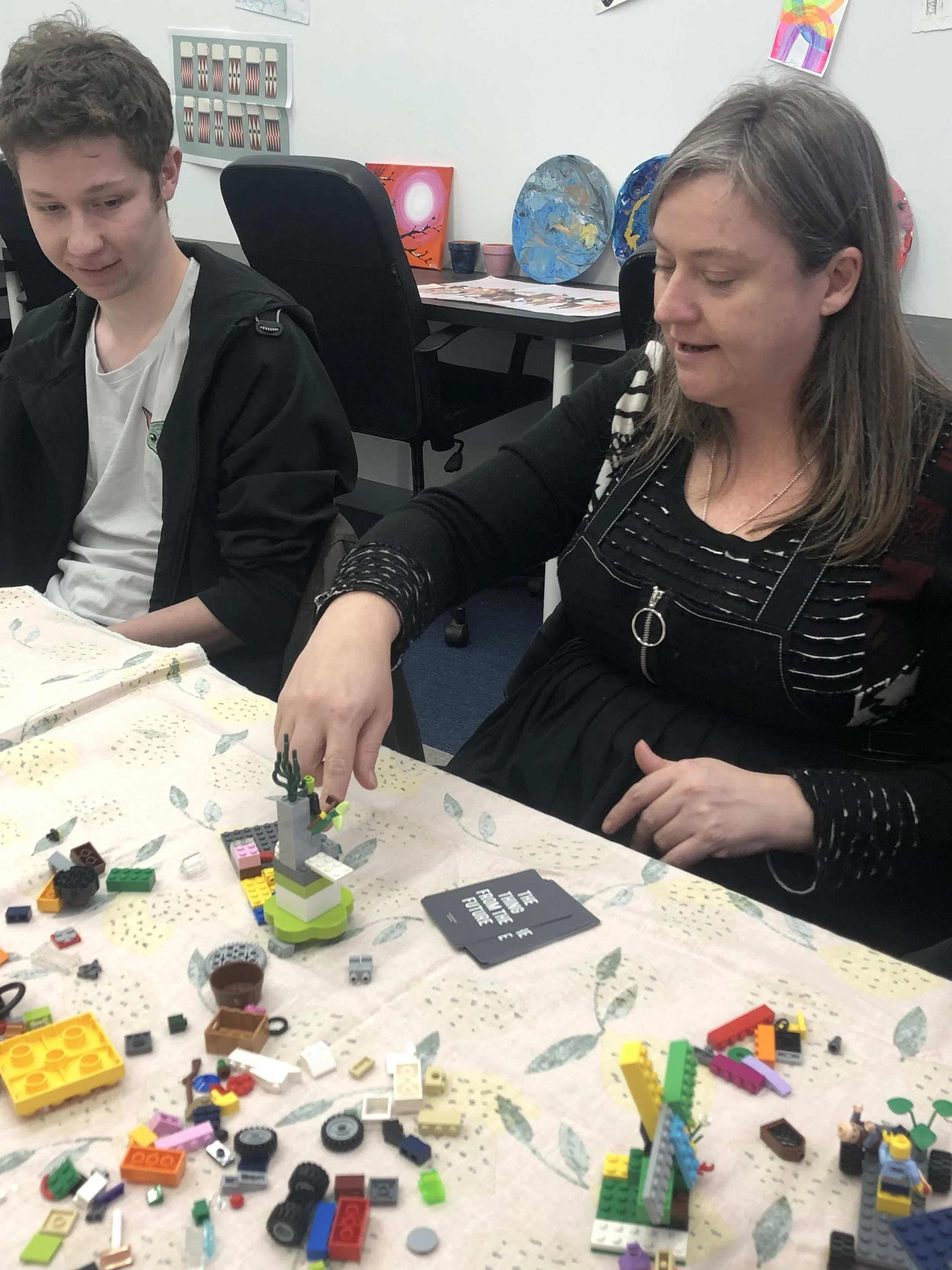 A woman and a teenage boy sitting at a table with LEGO pieces, with the woman placing a LEGO figure on a small LEGO structure.