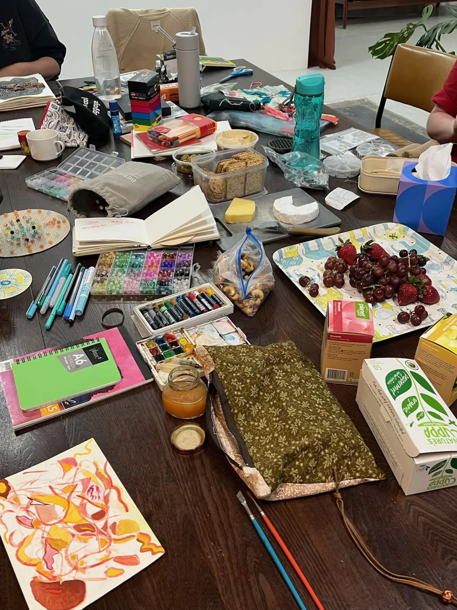 A table filled with art supplies, food, and drink items, including paint sets, pens, notebooks, cookies, strawberries, grapes, juice boxes, and various containers, with a few people seated around it.