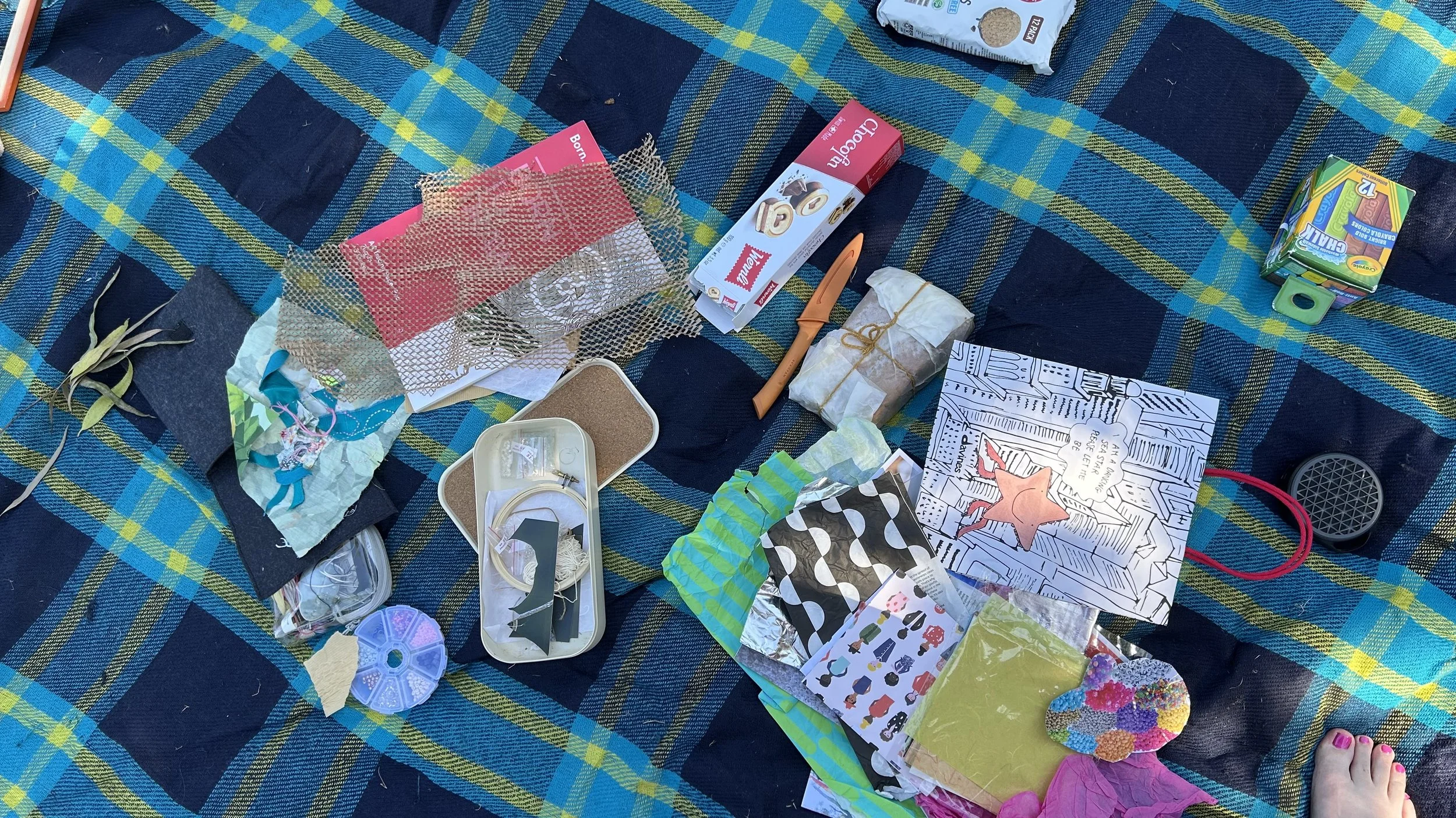 Assorted craft supplies and gift wrapping items spread on a blue plaid blanket, including paper, tape, scissors, fabric, stickers, and decorative objects.