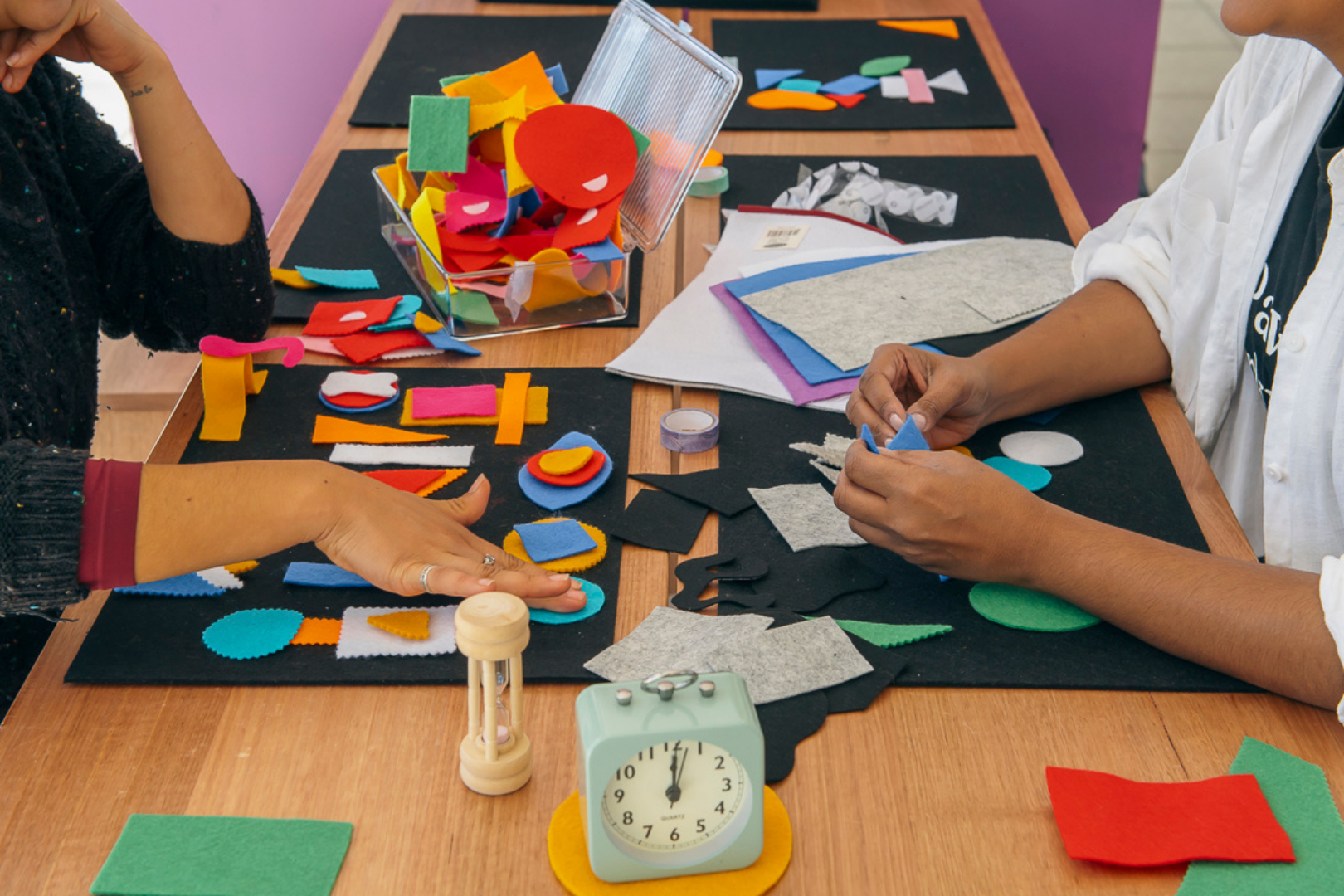 People engaging in a felt craft project at a wooden table, with colorful felt pieces, scissors, and a light green clock in the foreground.
