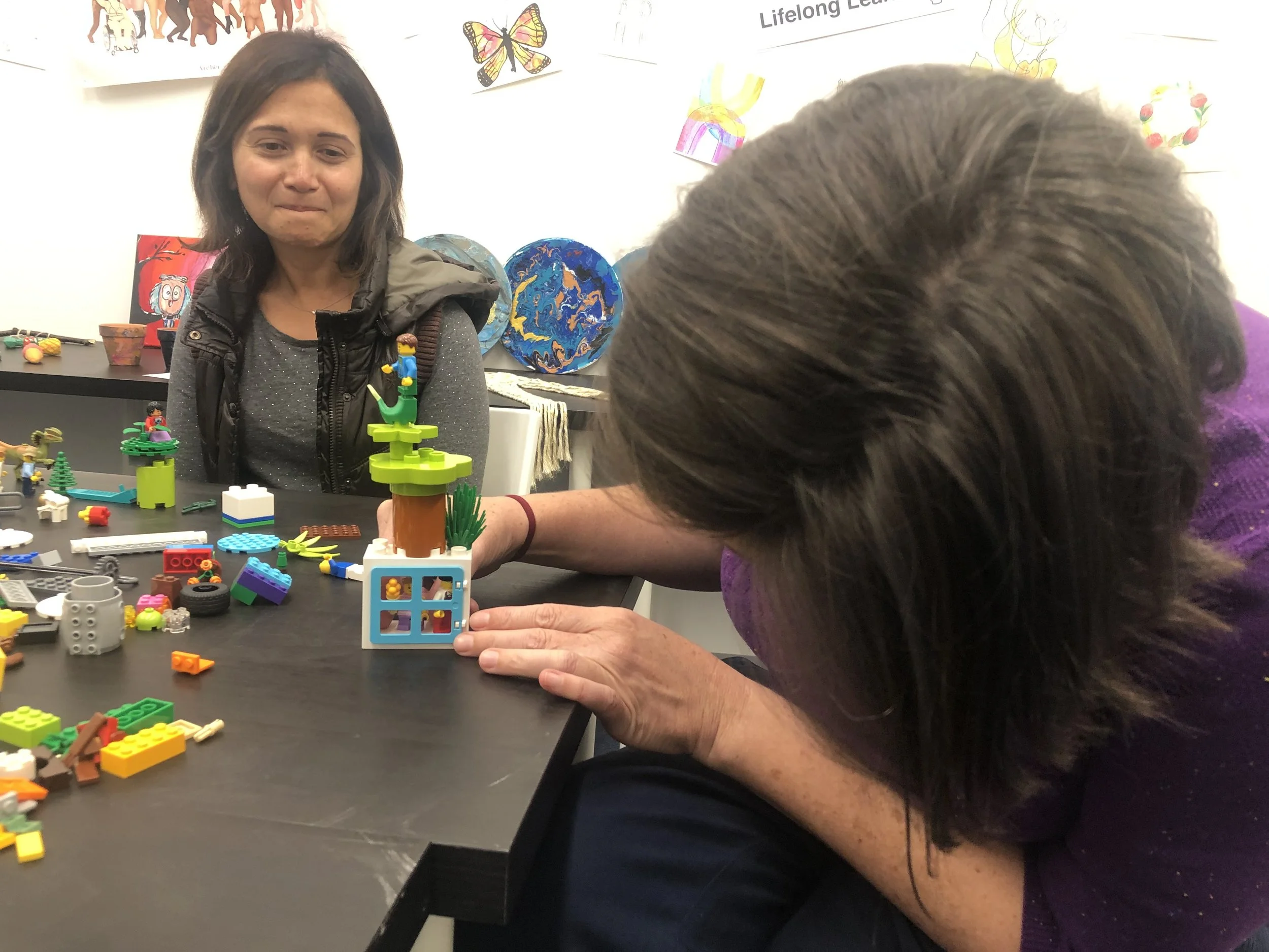 Two women are at a table filled with Lego bricks, working on building a Lego creation that appears to be a small tree with a figure on top. One woman is looking at the creation, while the other woman is leaning over and inspecting or adjusting the model. The background shows colorful artwork and posters on the wall.