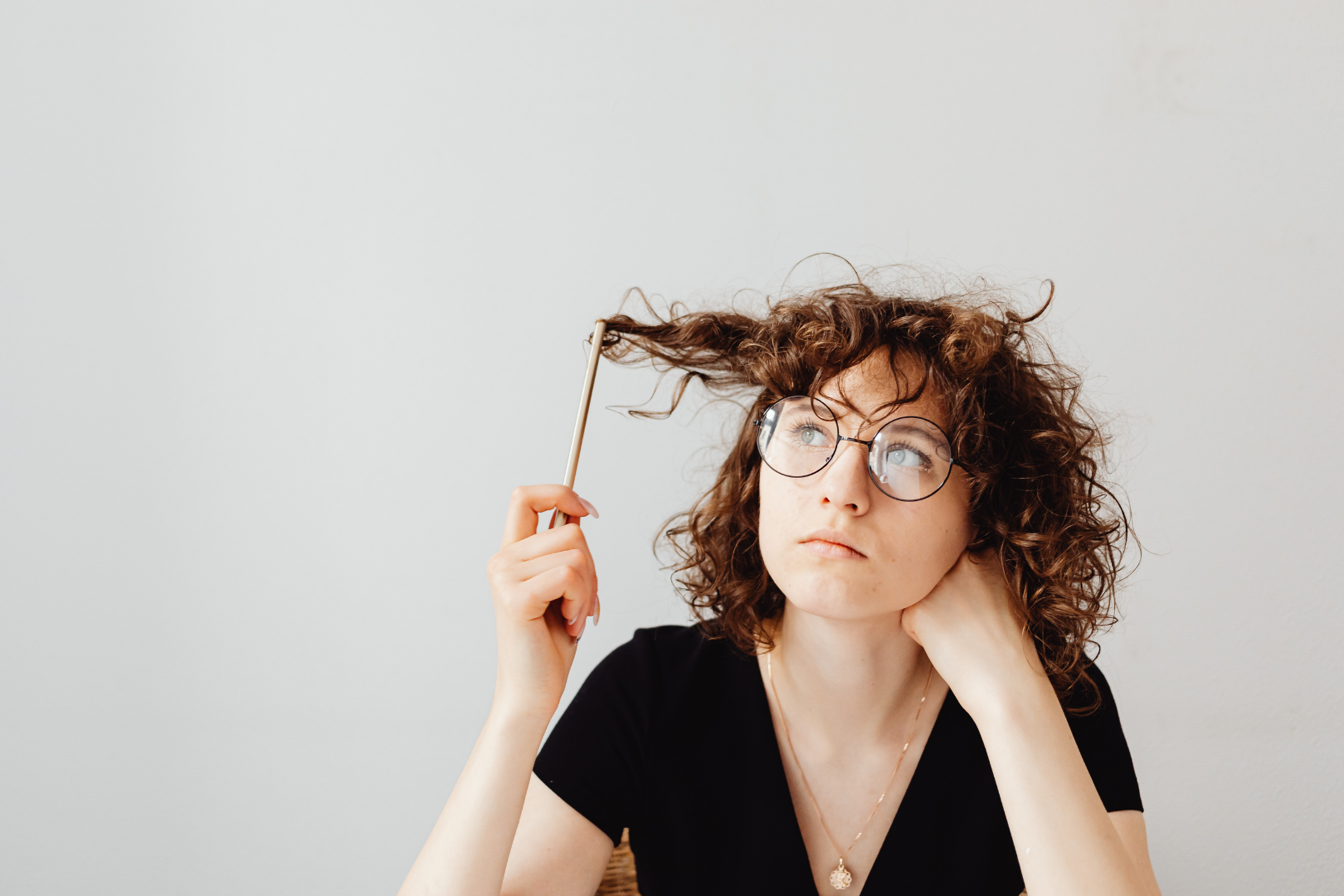 A youthful woman wearing glasses and a black top twirls her curly hair with a pencil while she stares into space