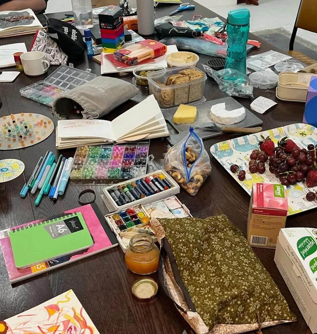 A photo of a table strewn with art materials, artworks, fruit, nuts, and cheese
