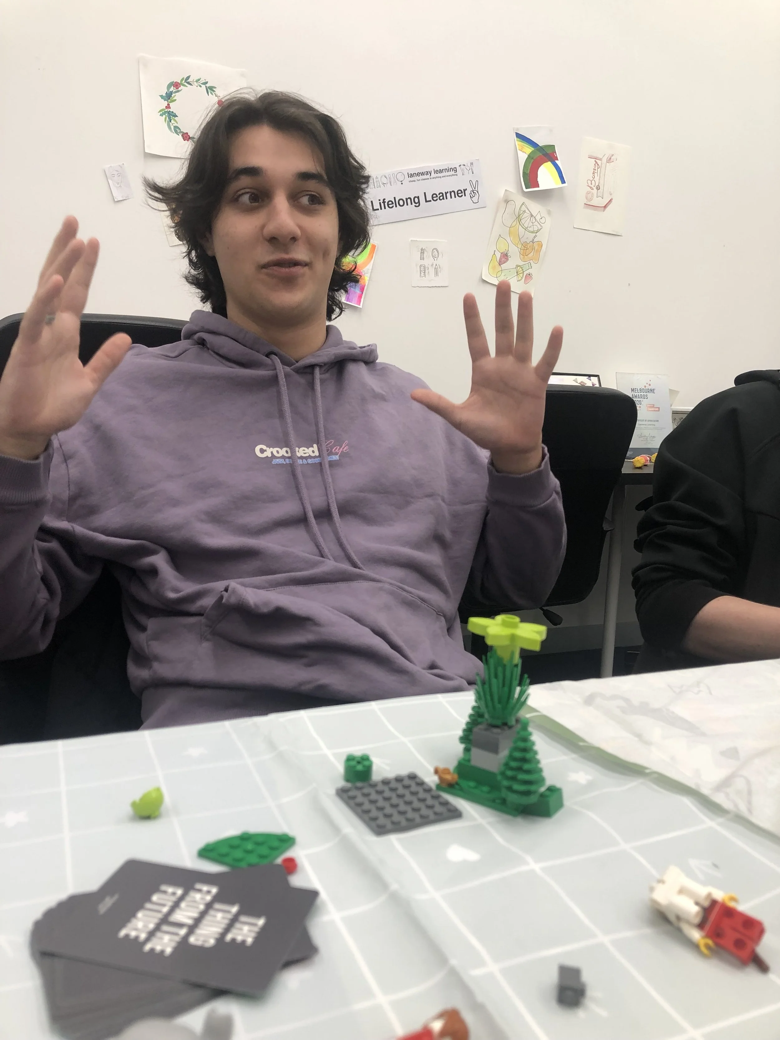 A young man with dark hair wearing a purple hoodie sitting at a table with Lego building blocks, partially built Lego trees, and some scattered pieces. In the background, there are colorful drawings and signs on the wall.