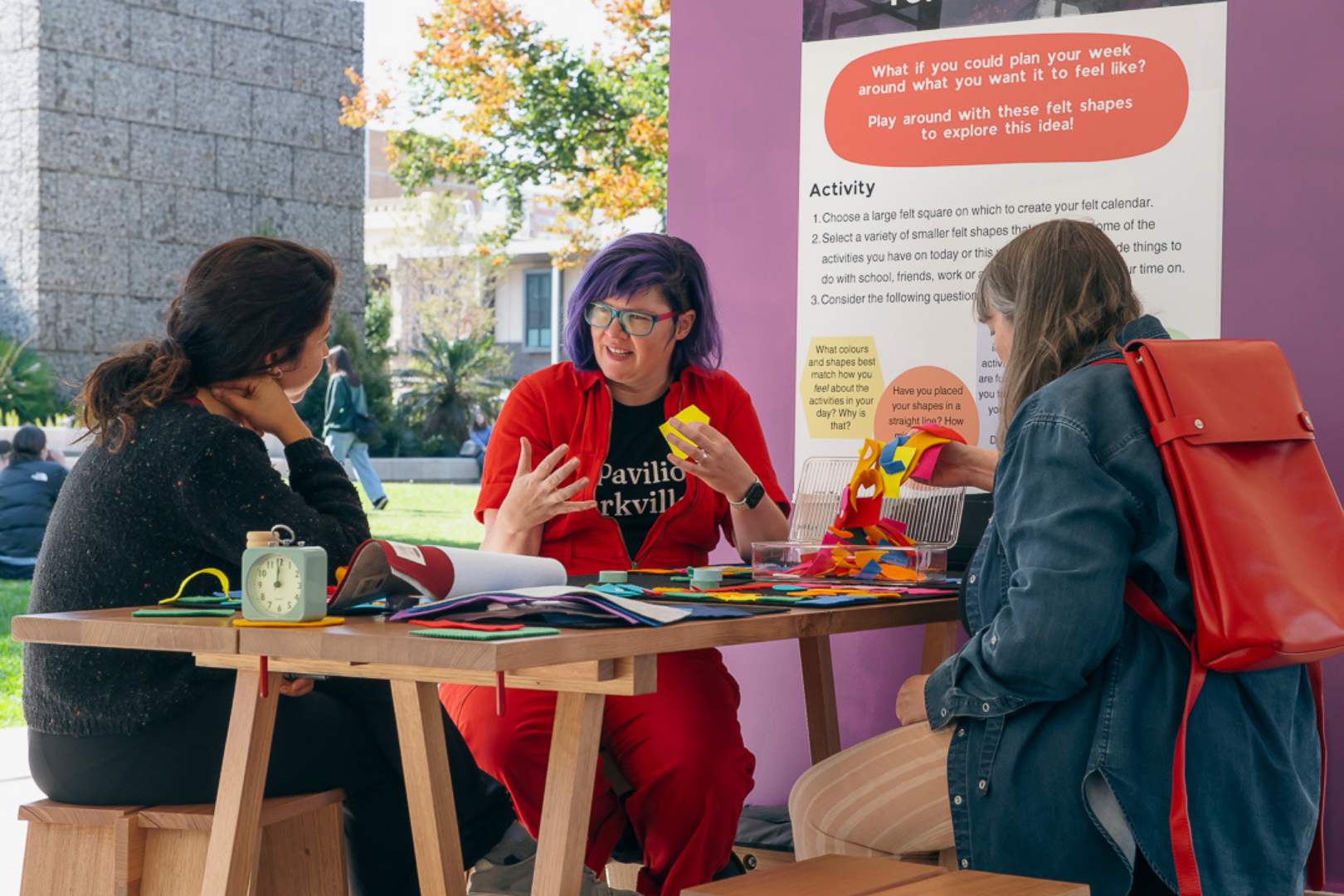 Three women sitting at a wooden table outdoors, engaging in a craft activity with felt shapes, in front of a purple display board with educational prompts and questions related to felt shapes and activities, some trees, people, and a stone building in the background.