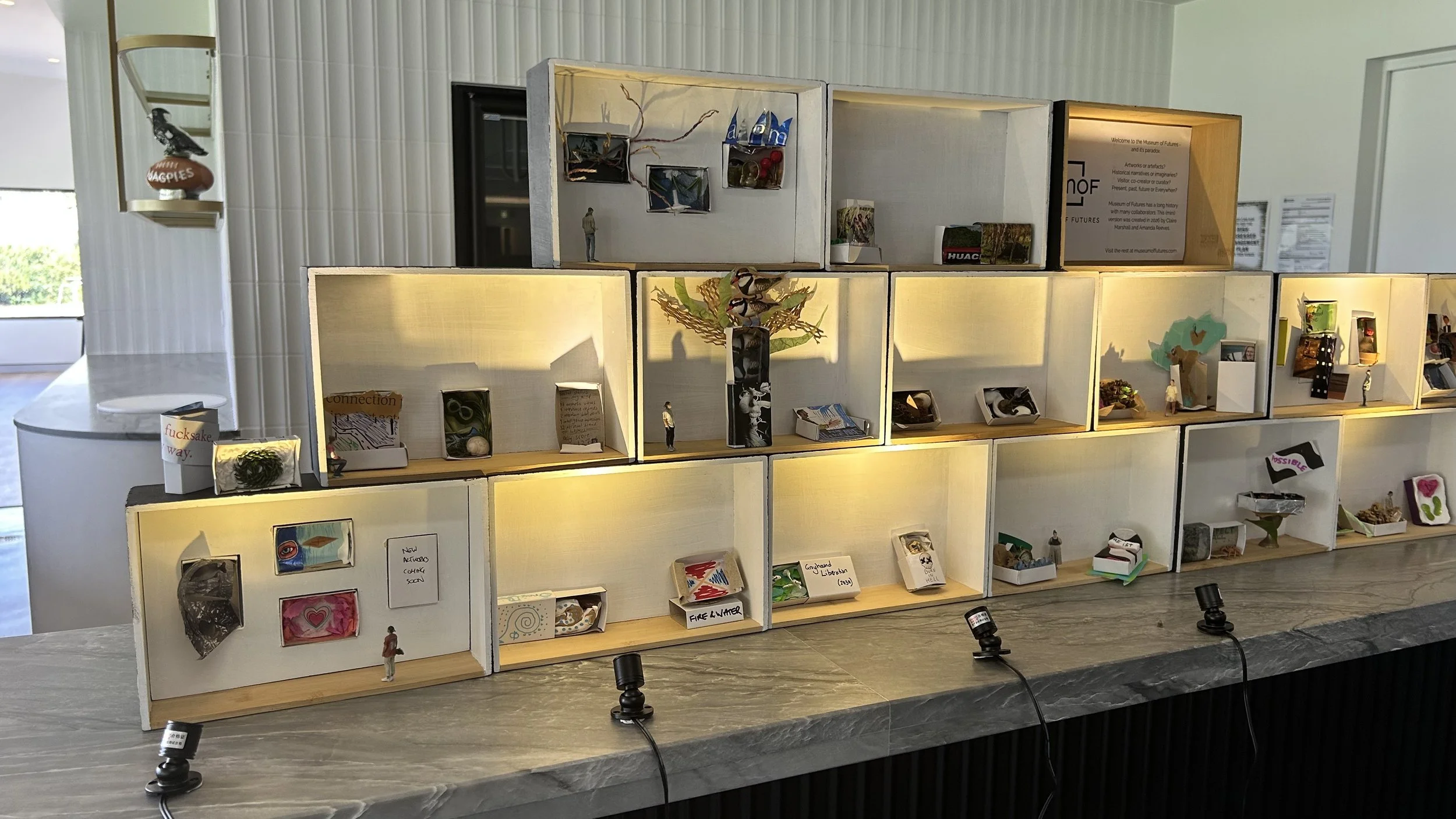 Miniature dioramas on a table depicting various scenes and objects in illuminated display boxes, with a white wall and window in the background.
