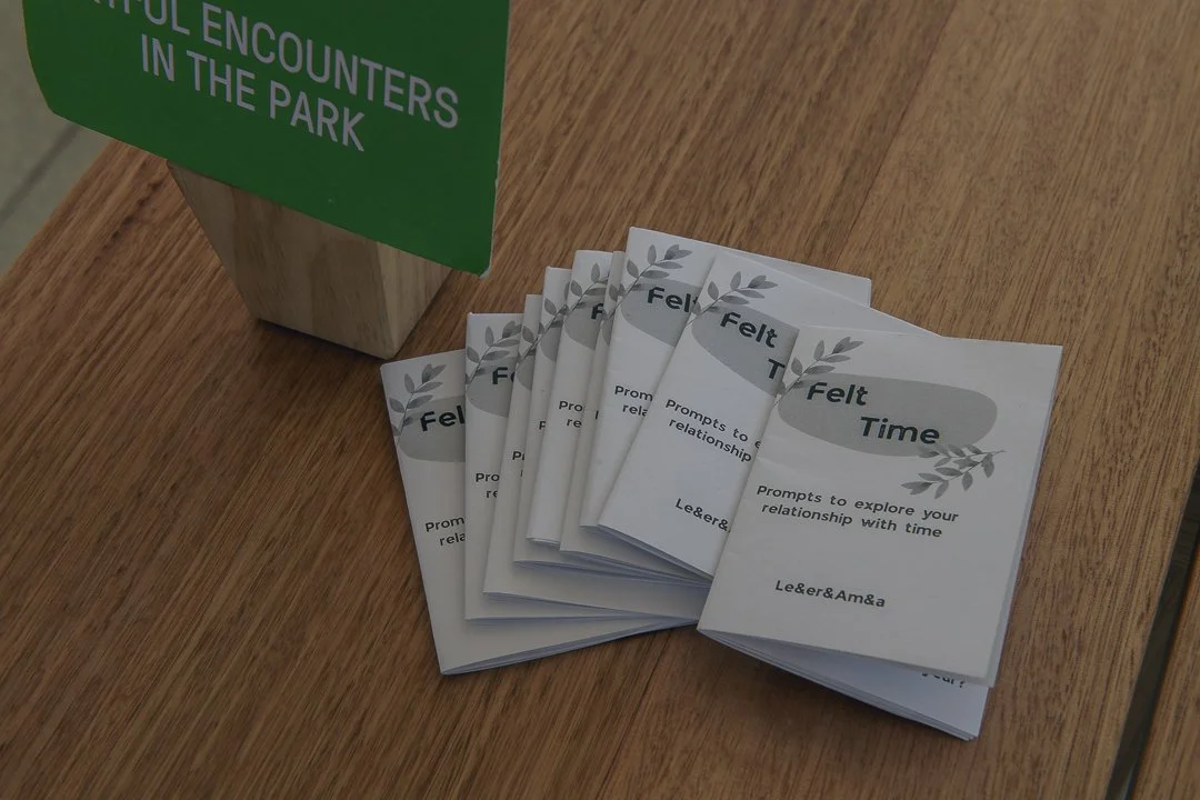 Pamphlets titled 'Felt Time' promoting exploring relationships with time, placed on a wooden table near a green sign that says 'FUL ENCOUNTERS IN THE PARK'.
