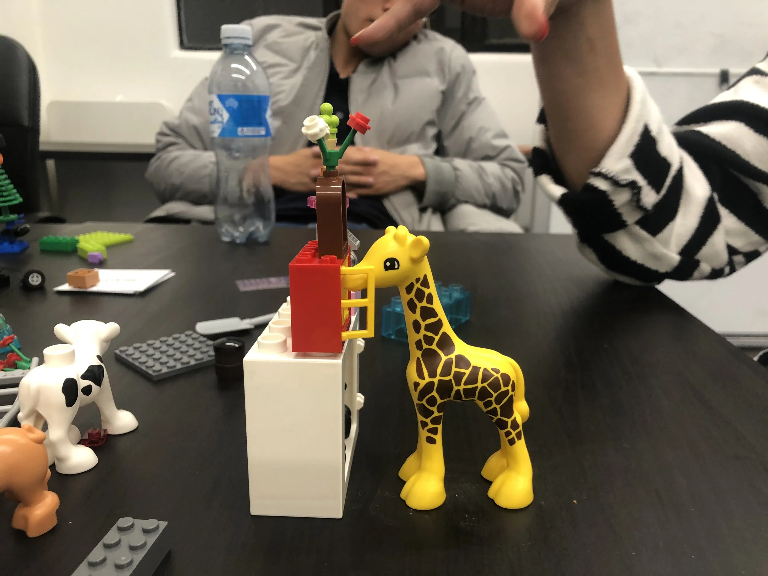 Colorful LEGO animal figures, including a giraffe, on a black table surrounded by other LEGO pieces and a water bottle; people seated in the background.