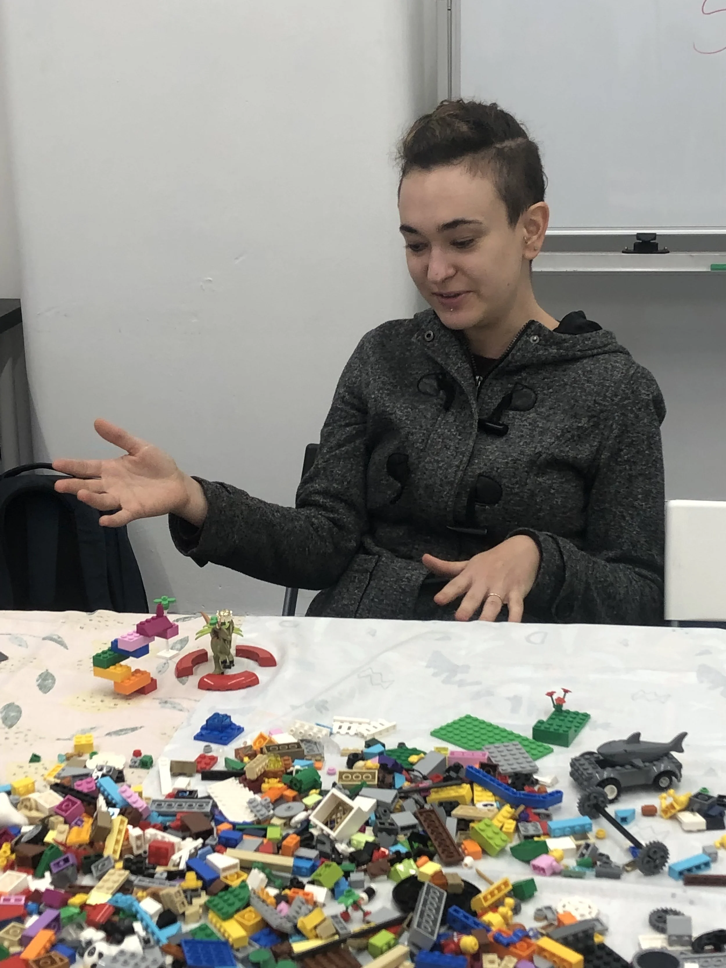 A woman sitting at a table with a large pile of colorful LEGO bricks and figures, gesturing with her right hand, in a room with a whiteboard in the background.