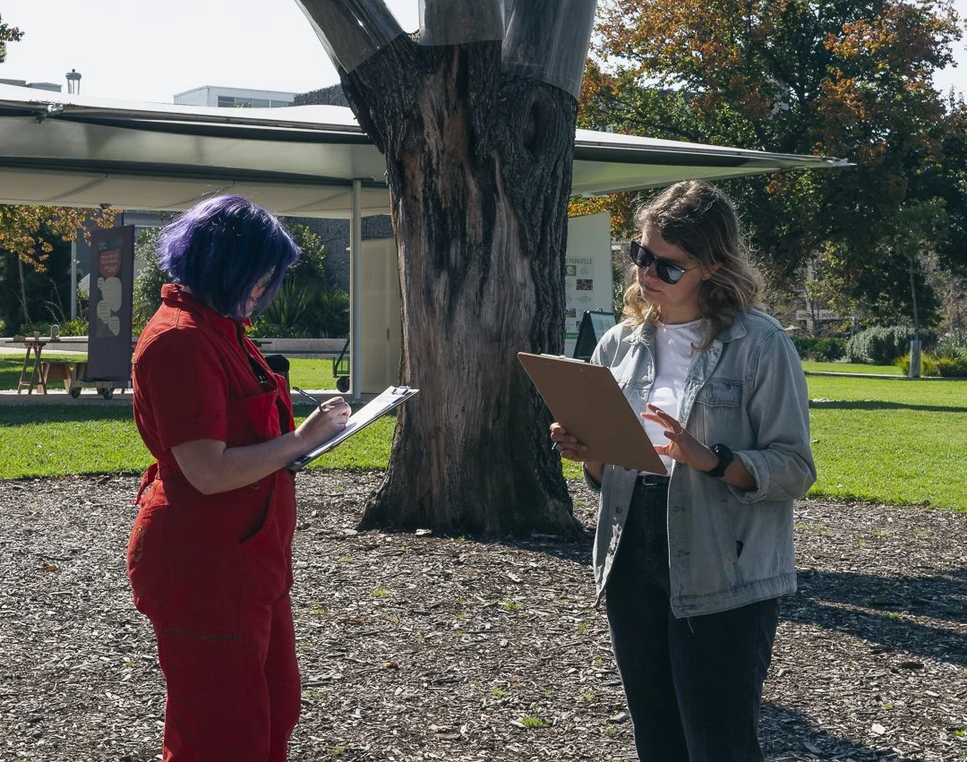 Two women standing outside under a large tree, looking at clipboards, one taking notes and the other speaking, with a park and a building in the background during daytime.