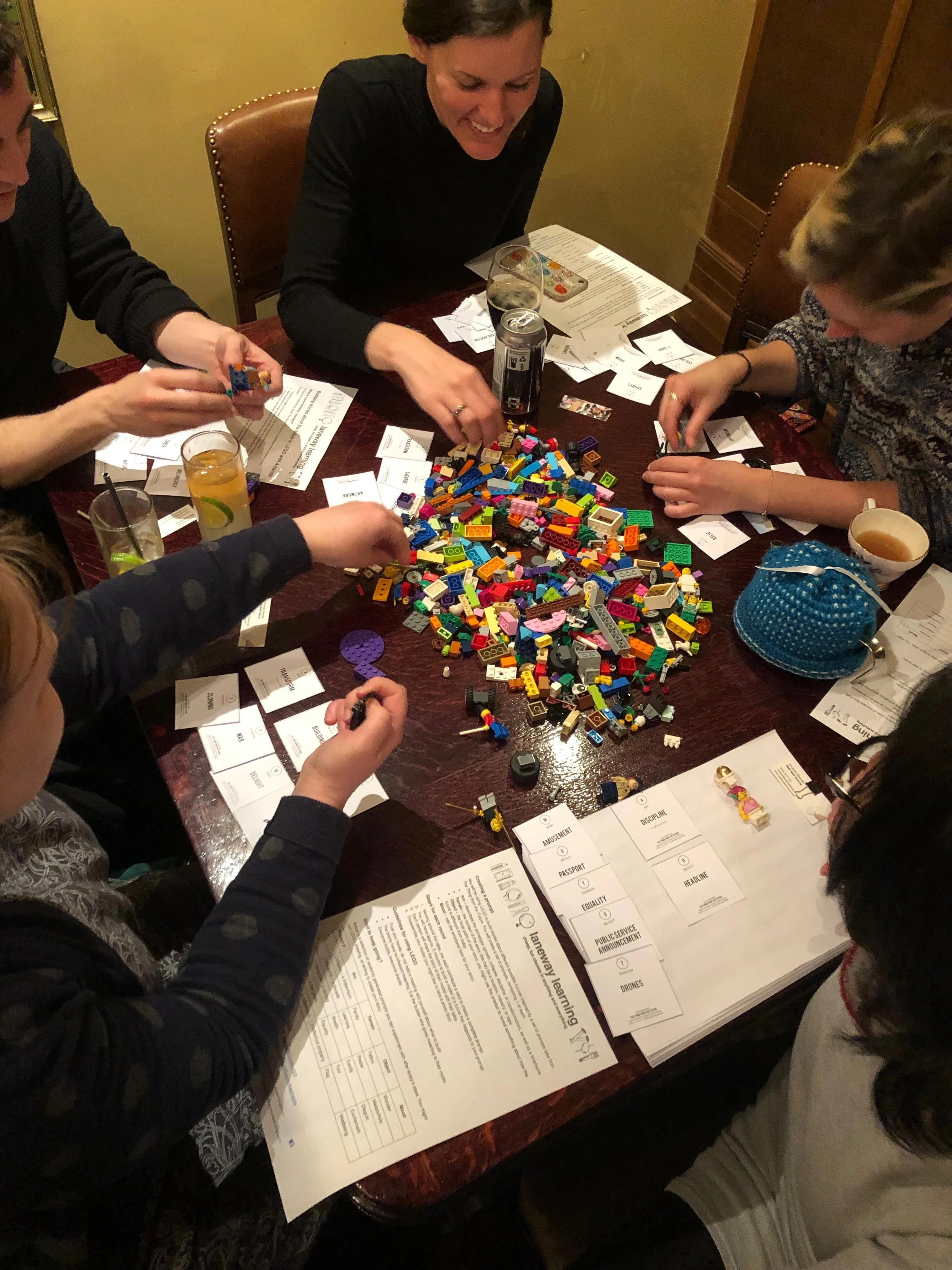 Group of people sitting around a table playing with LEGO bricks, with papers and drinks on the table.