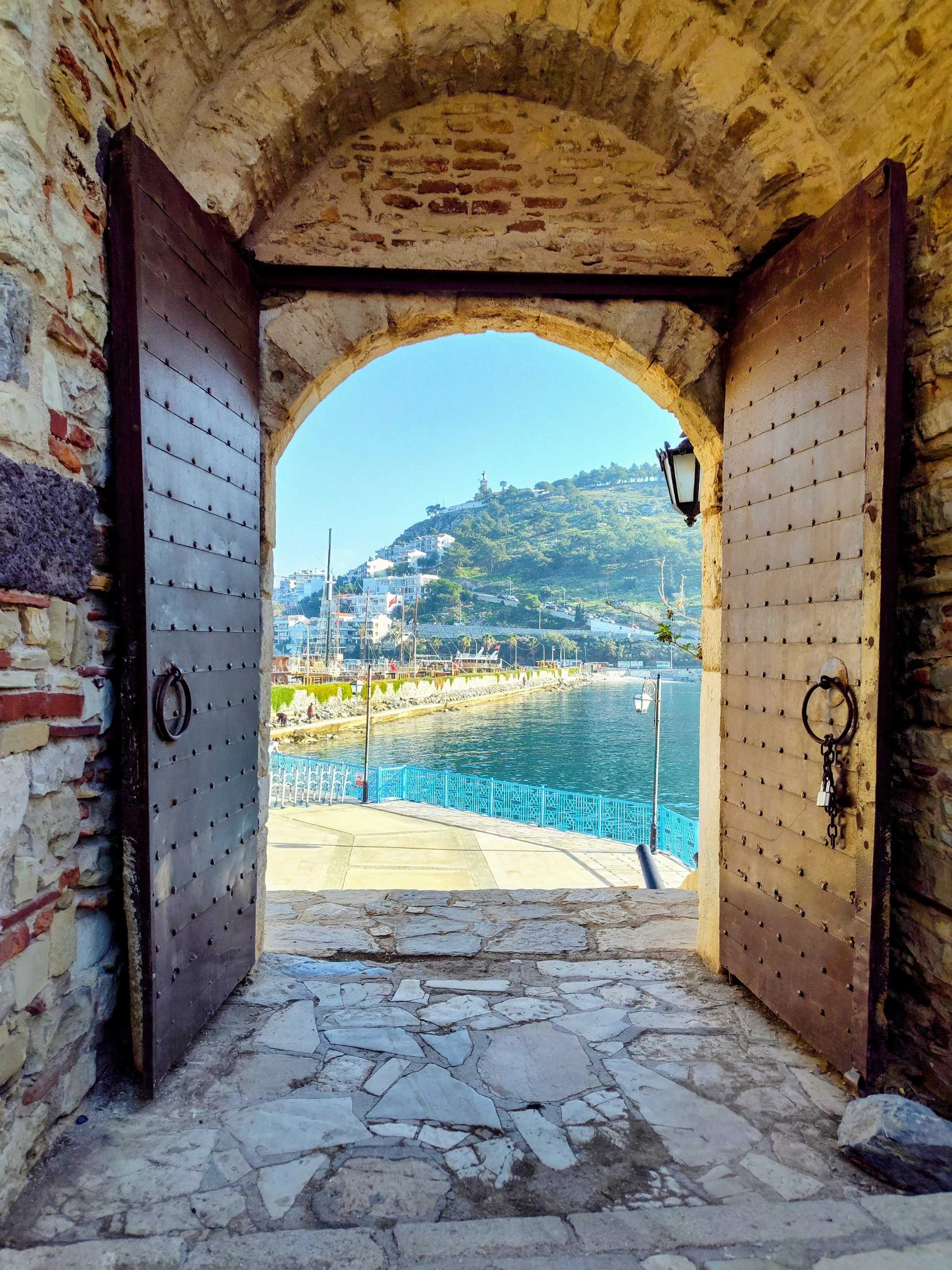 Original photography of the historic fortress gates in Kusadasi, Turkey, overlooking the Aegean Sea.  The open metal doors frame a vibrant view of white hillside villas, sailboat masts, and the deep blue Mediterranean water.