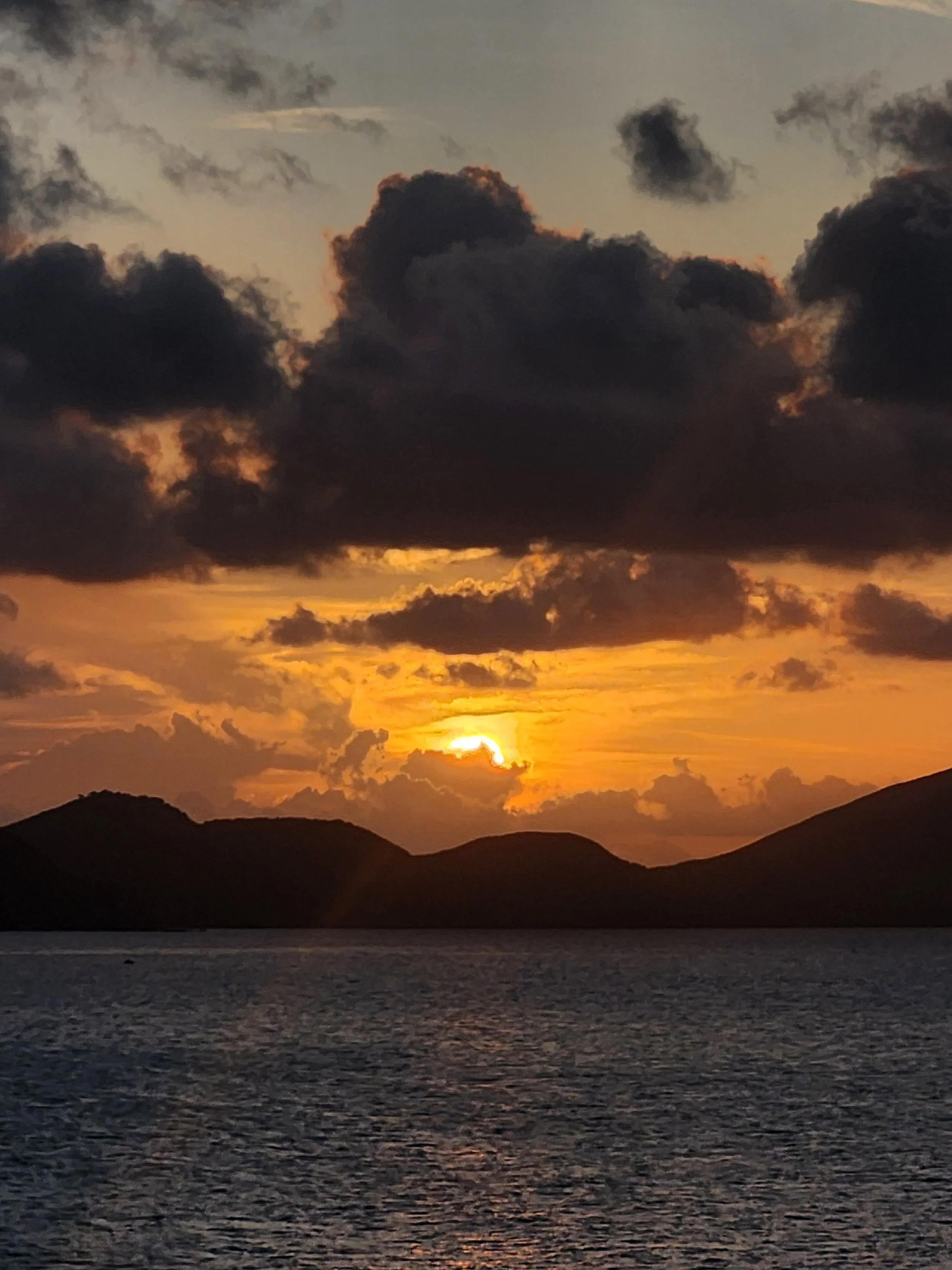 A vibrant orange sunset over the ocean with a silhouetted tropical island and clouds reflecting on the water, captured during a Sunrise Travel luxury cruise.