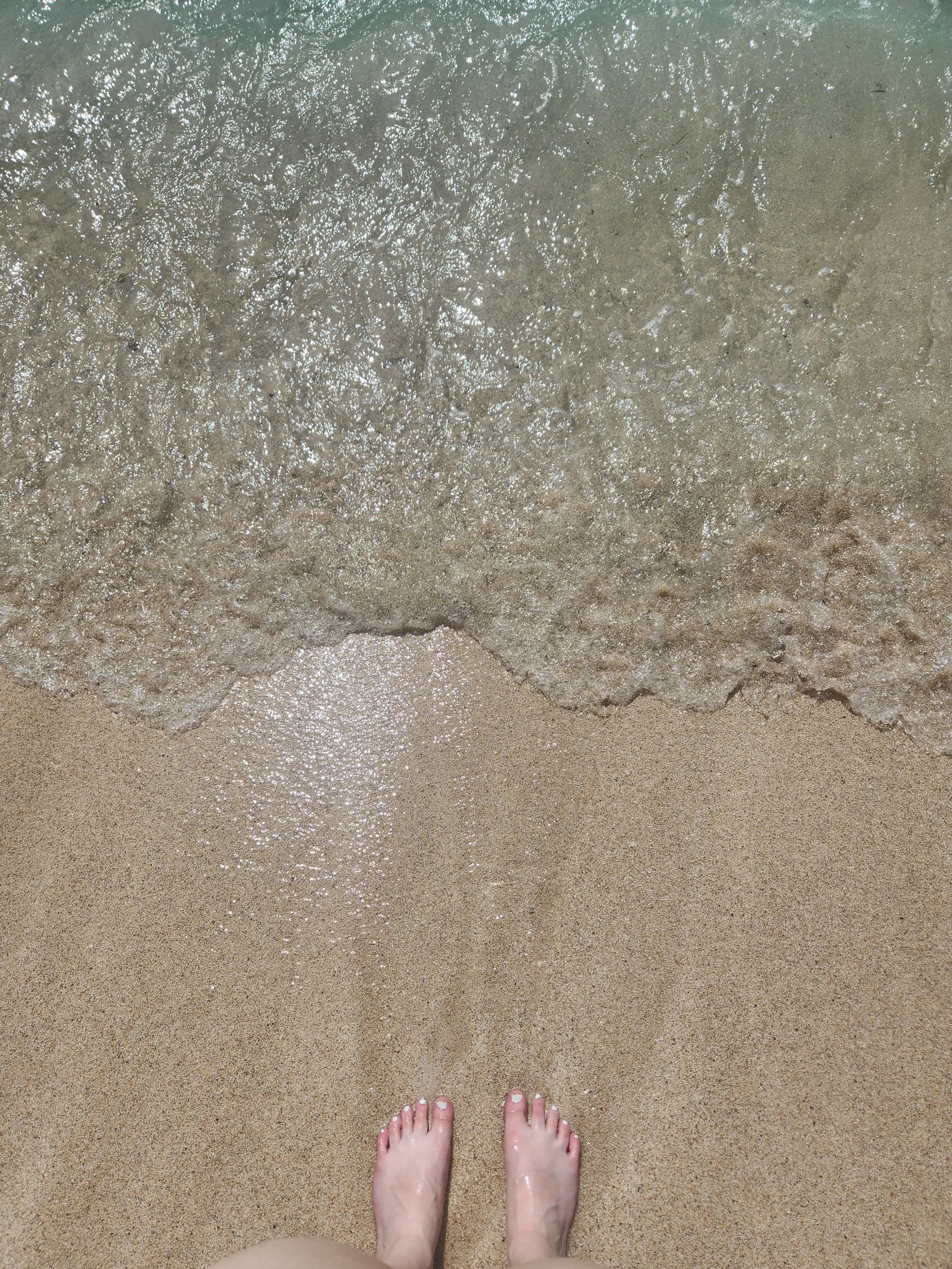 A first-person perspective of feet in soft warm sand with clear blue ocean water, showing the firsthand Caribbean travel experience provided by Sunrise Travel