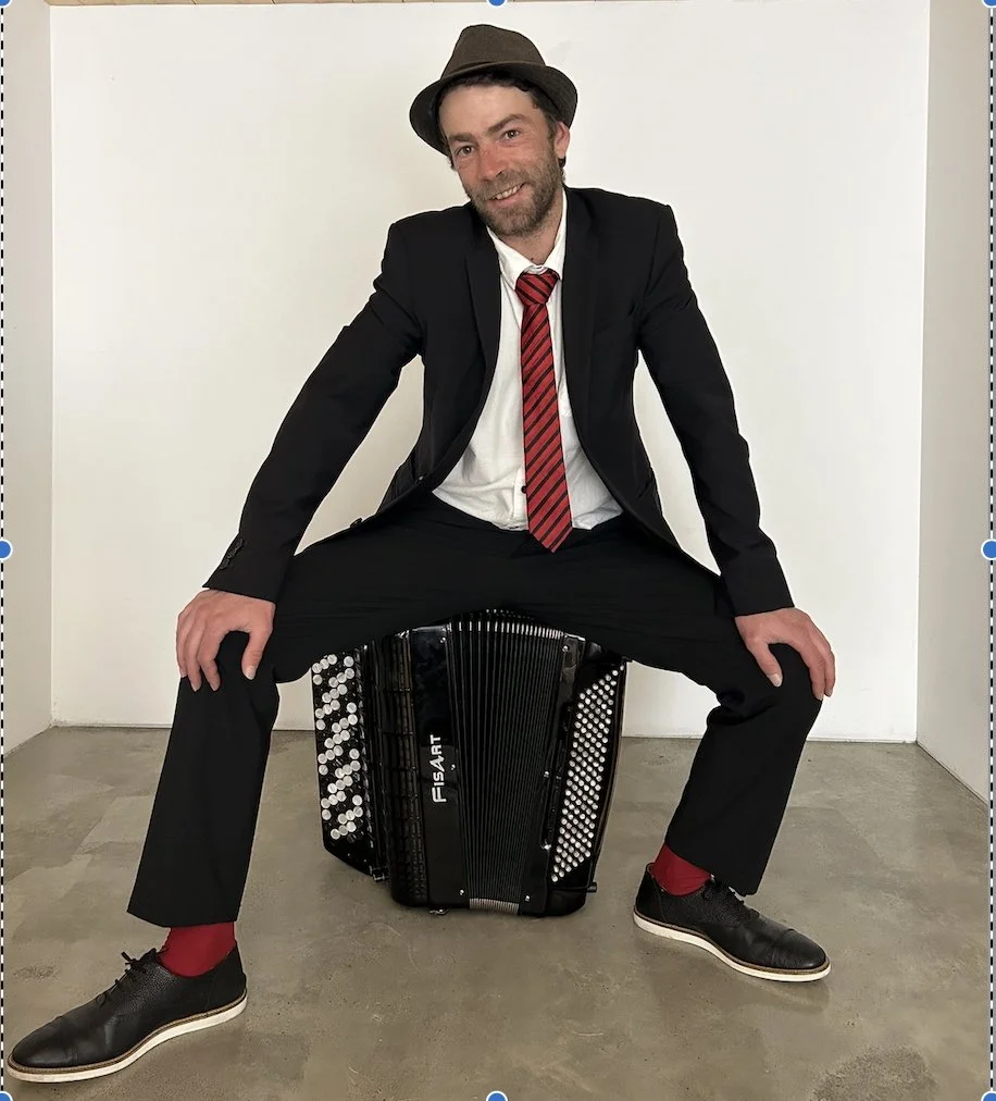 Un homme habillé en costume noir, chemise blanche, cravate rouge, chapeau gris, assis sur un accordéon.