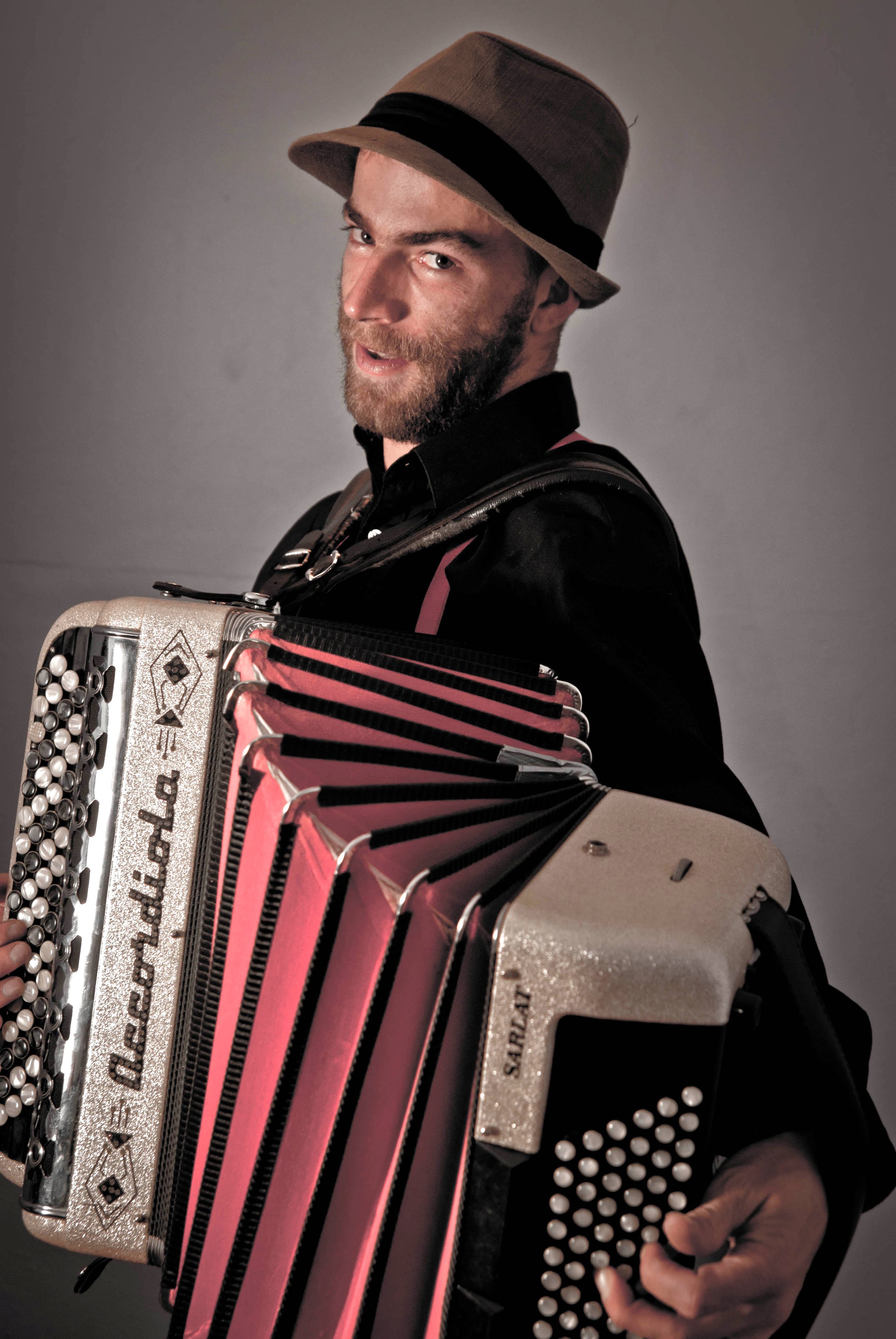 Un homme avec une barbe porte un chapeau gris et une chemise noire, il joue de l'accordéon avec des accents roses, sur un fond gris.