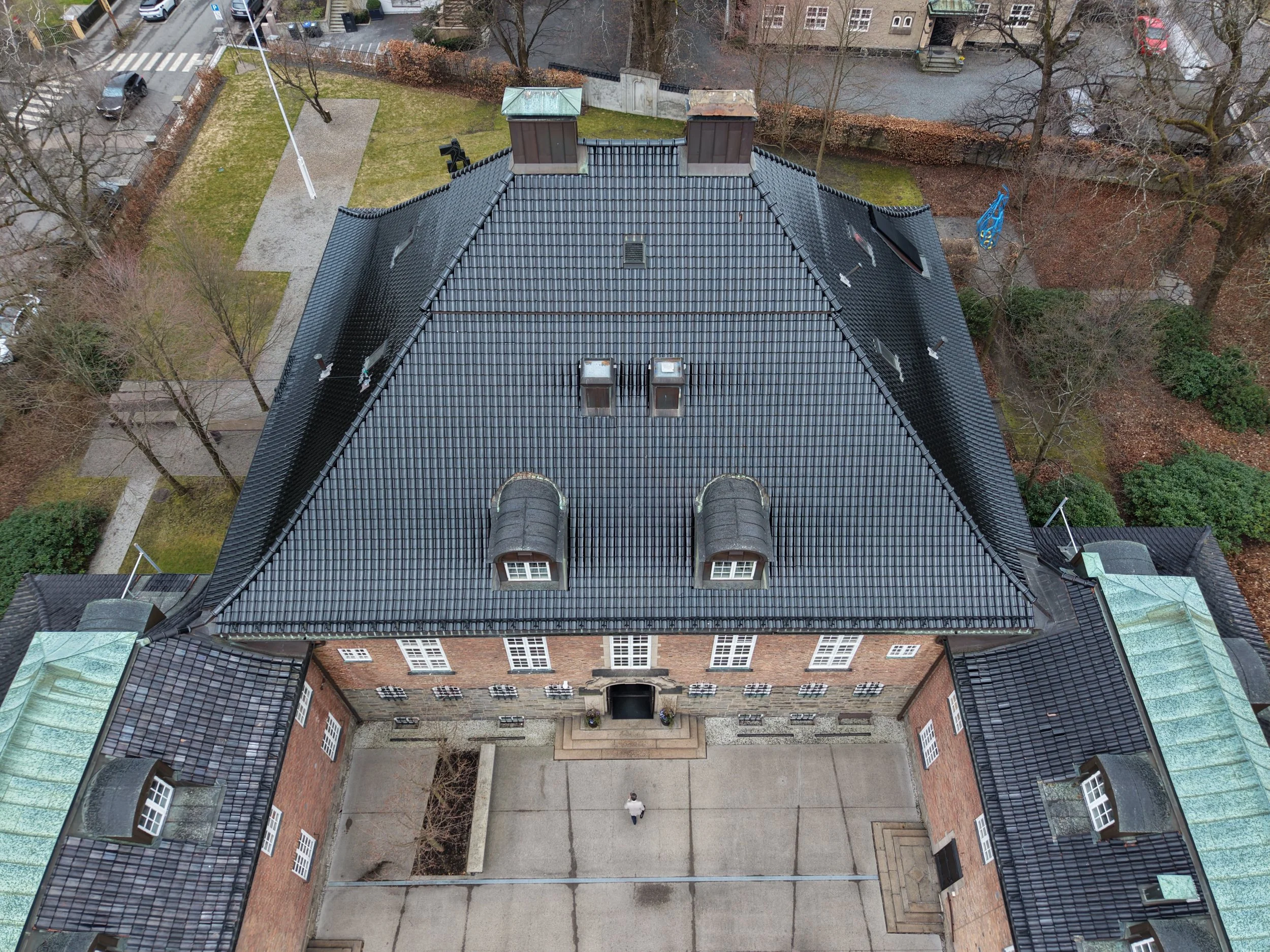 Utsikt over taket til en stor, eldre bygning med svarte taksteiner, omgitt av trær og en liten hage med gress, vegg, gangsti og noen personer under. Hovedinngang med trapp er synlig foran bygningen. Dronefoto