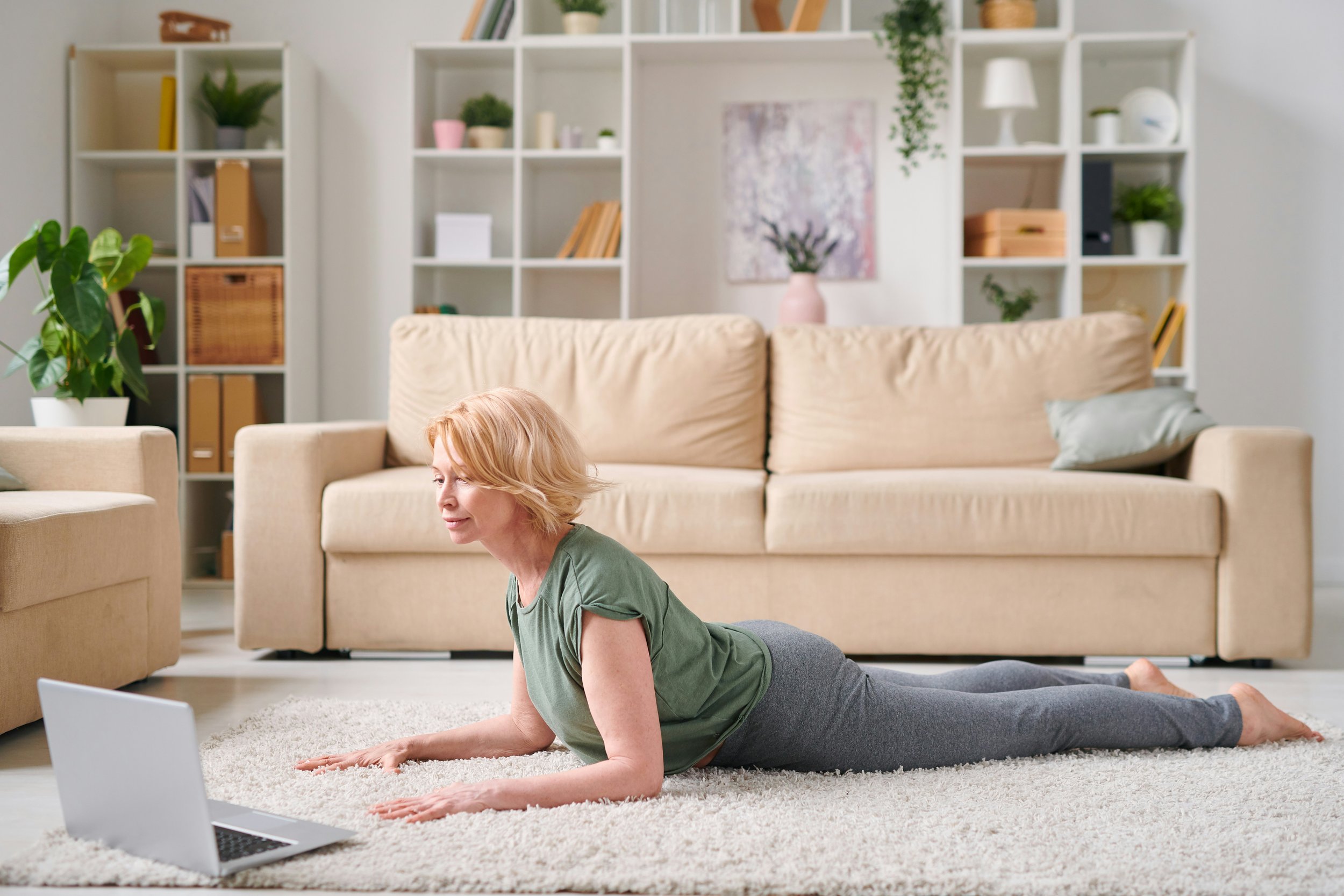 A woman practicing yoga in her living room with a laptop in front of her, on a light-colored rug. She is in the upward dog pose, wearing a green t-shirt and gray pants, in front of a beige sofa and white shelving filled with plants and decorative items.