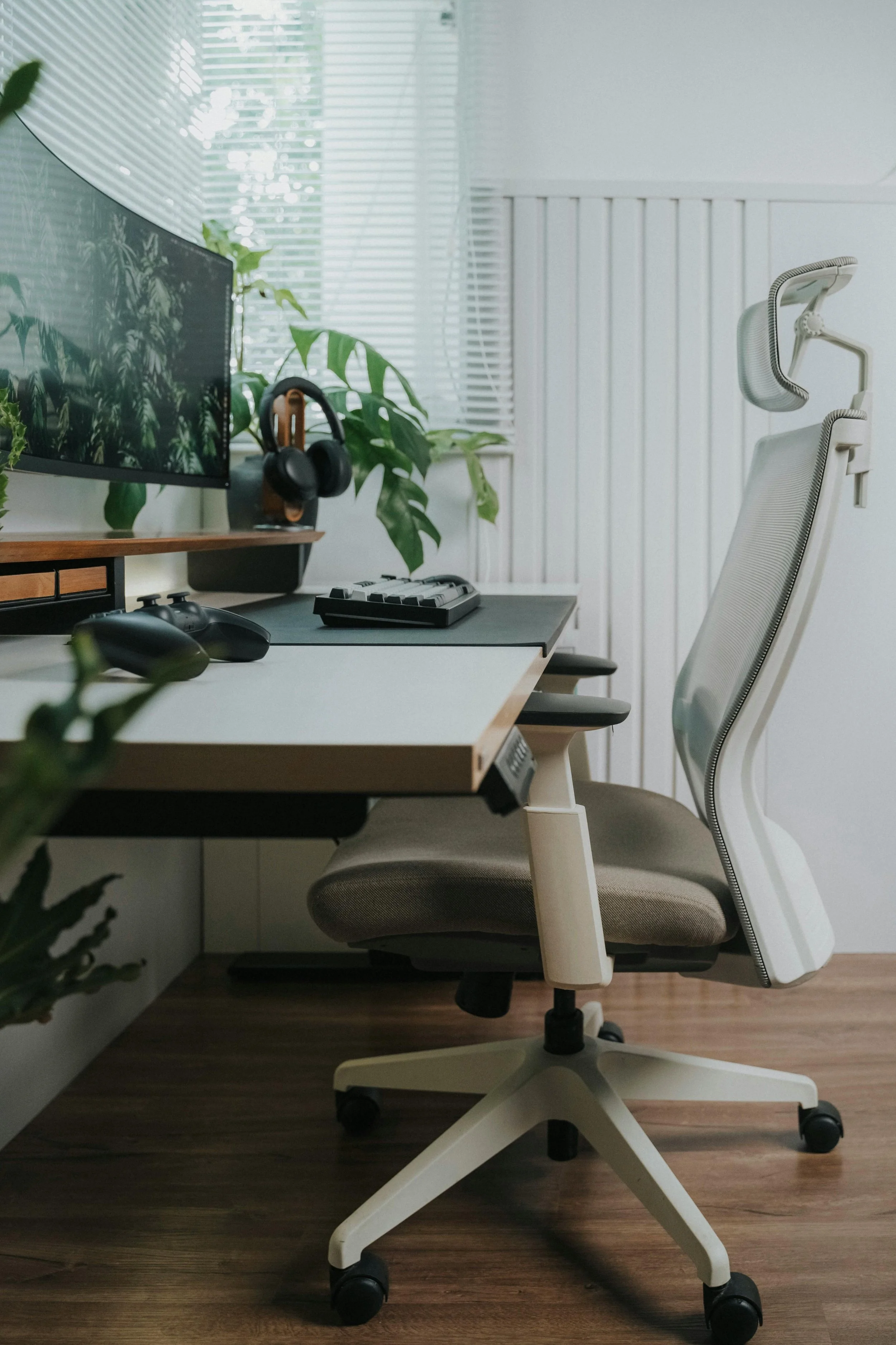 Modern home office setup with ergonomic white office chair, desk with computer monitor, keyboard, mouse, and headphones, along with potted plants and window blinds.