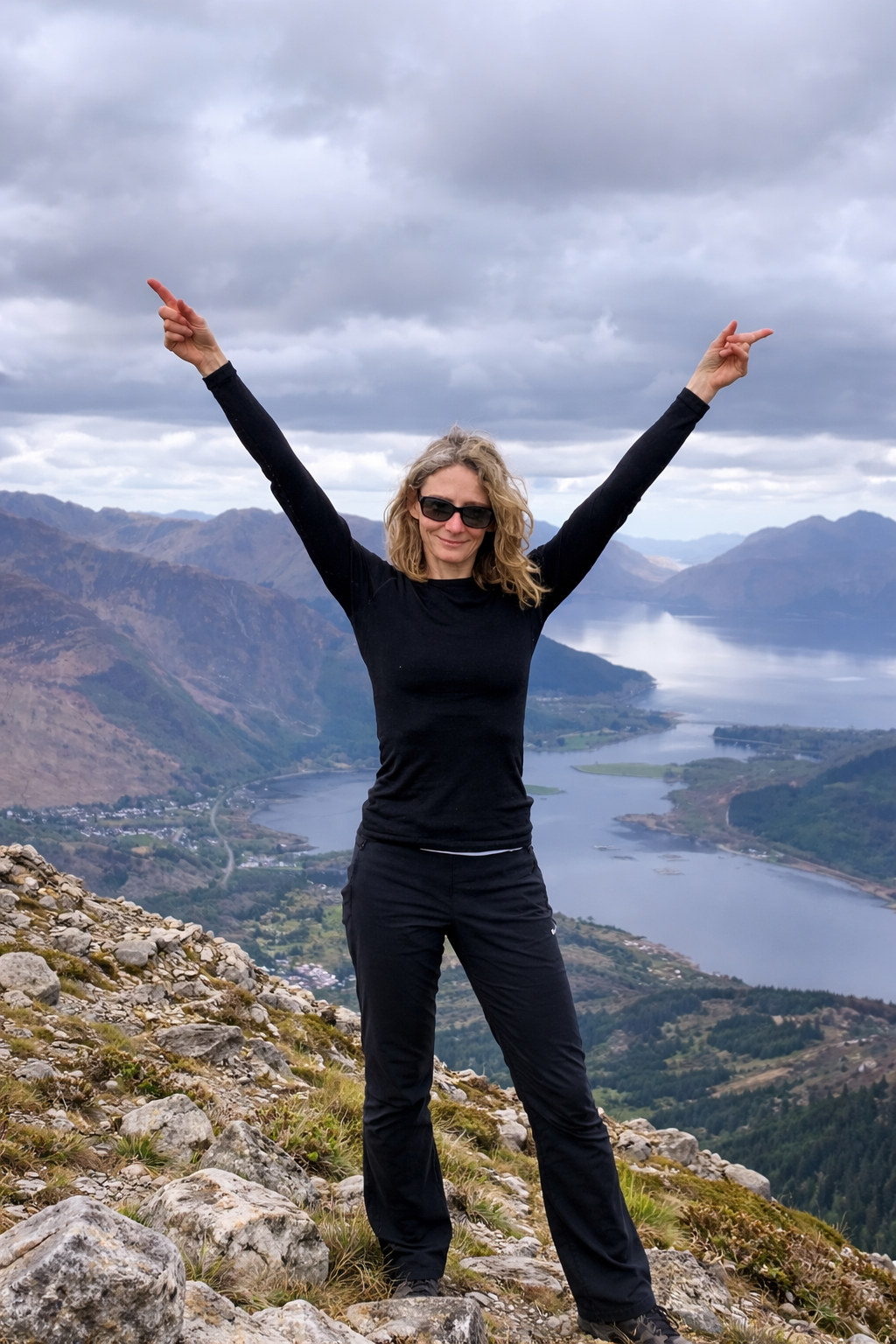 Woman in black outdoor clothing and sunglasses standing on rocky hillside with arms raised, overlooking a lake and mountain range under cloudy sky.