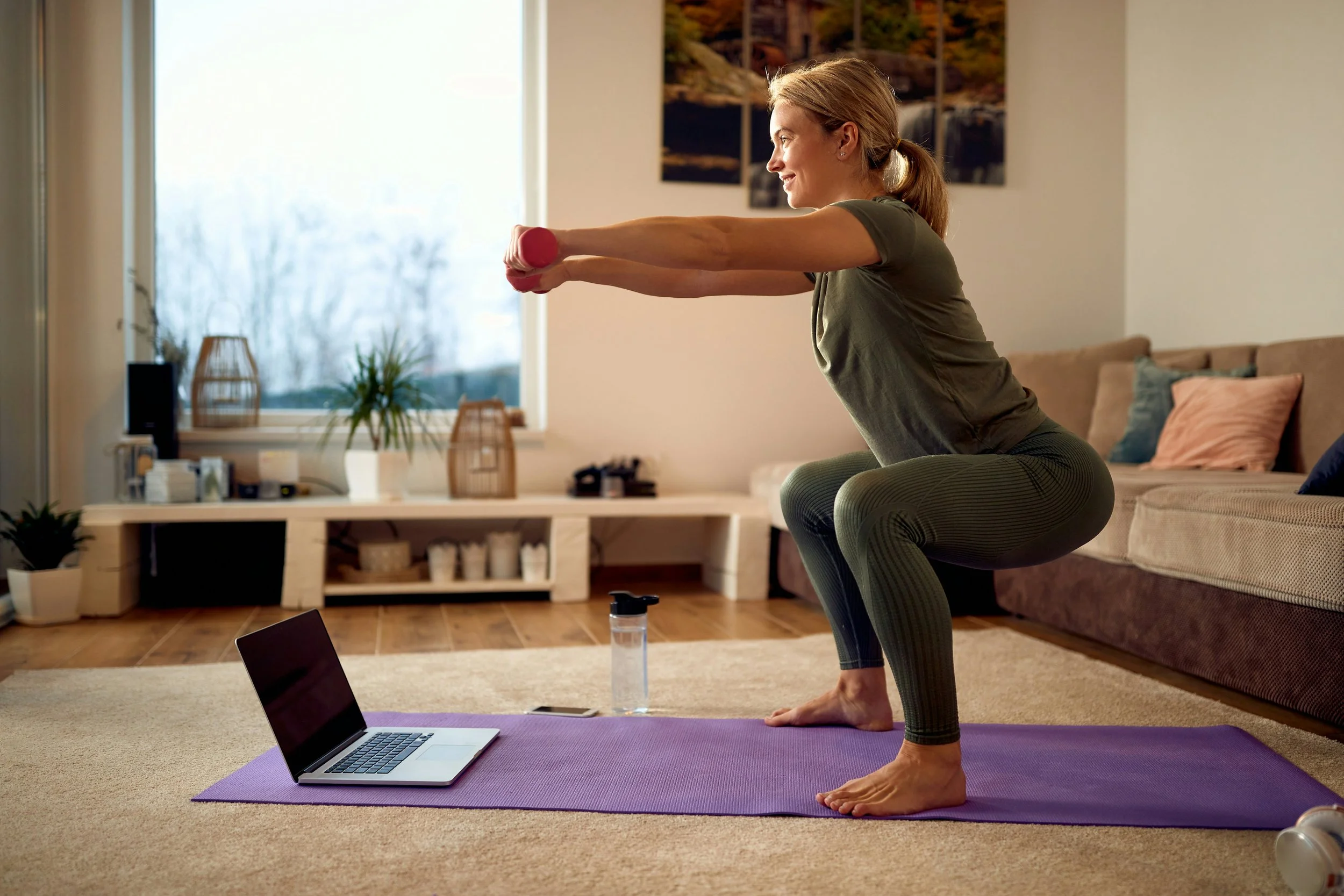 Woman practicing yoga outdoors in a forested area, sitting cross-legged on a mat with her hands resting on her knees, wearing a light gray tank top and black leggings.