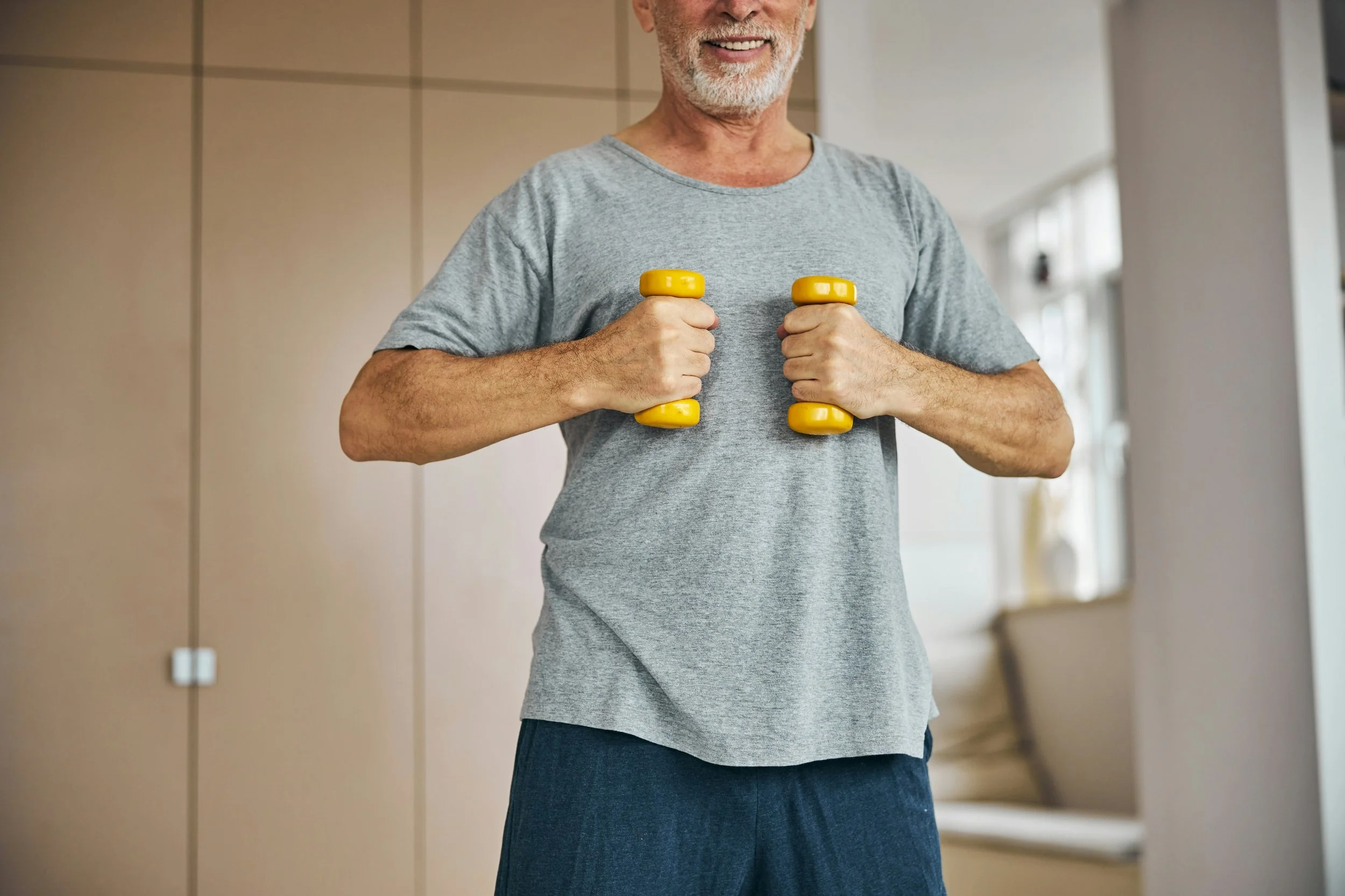 A man exercising indoors, holding yellow dumbbells close to his chest and smiling.