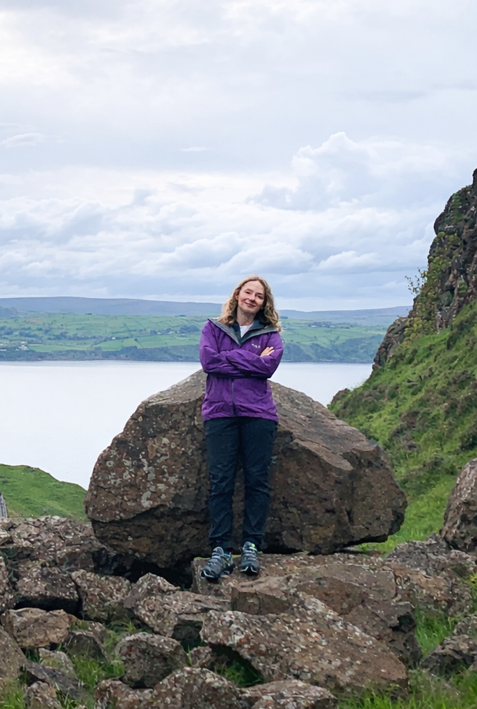 Woman standing on rocks near a body of water with hills and cloudy sky in background.