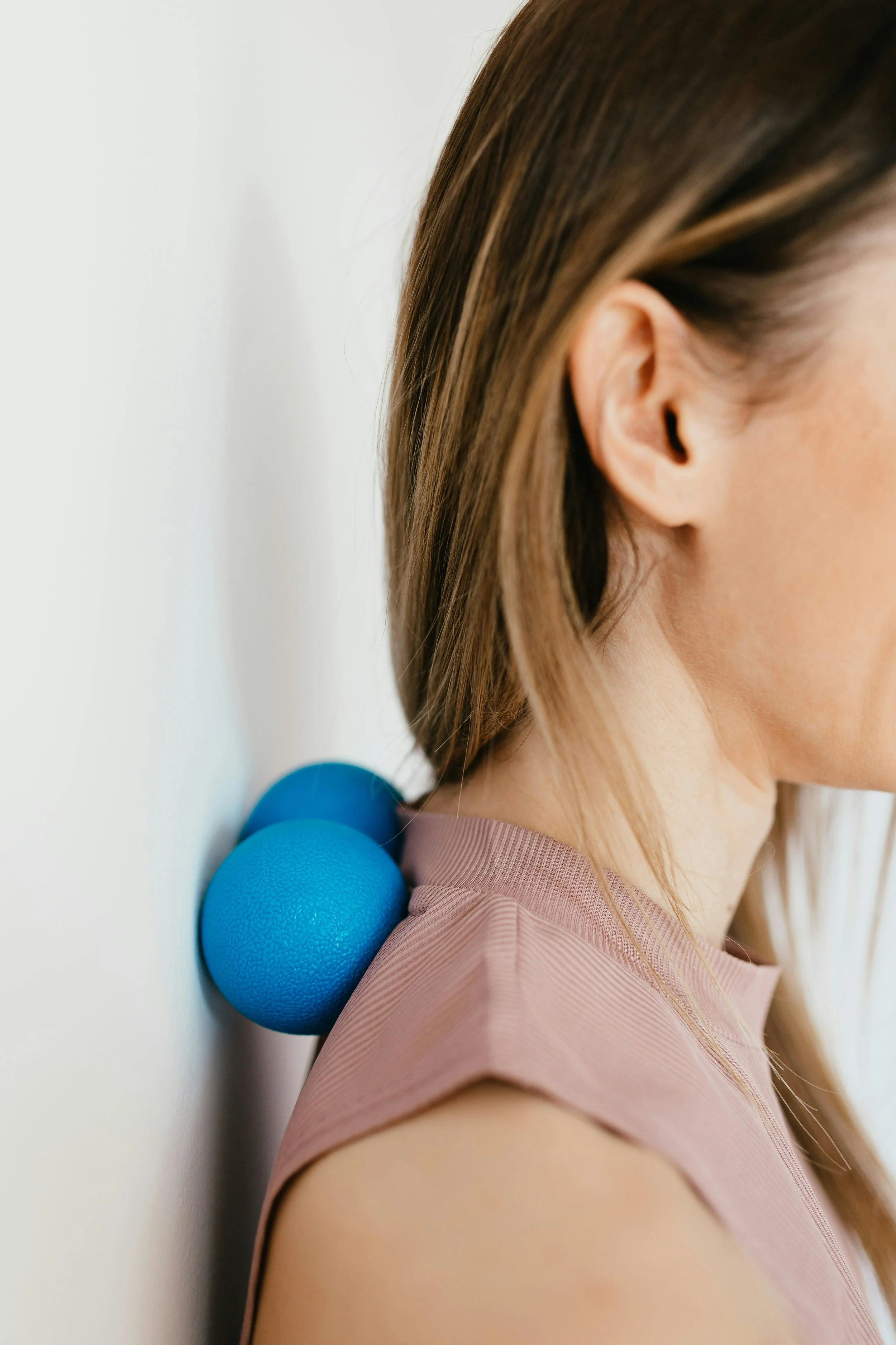 A woman leaning against a white wall with two blue textured massage balls resting on her shoulder.